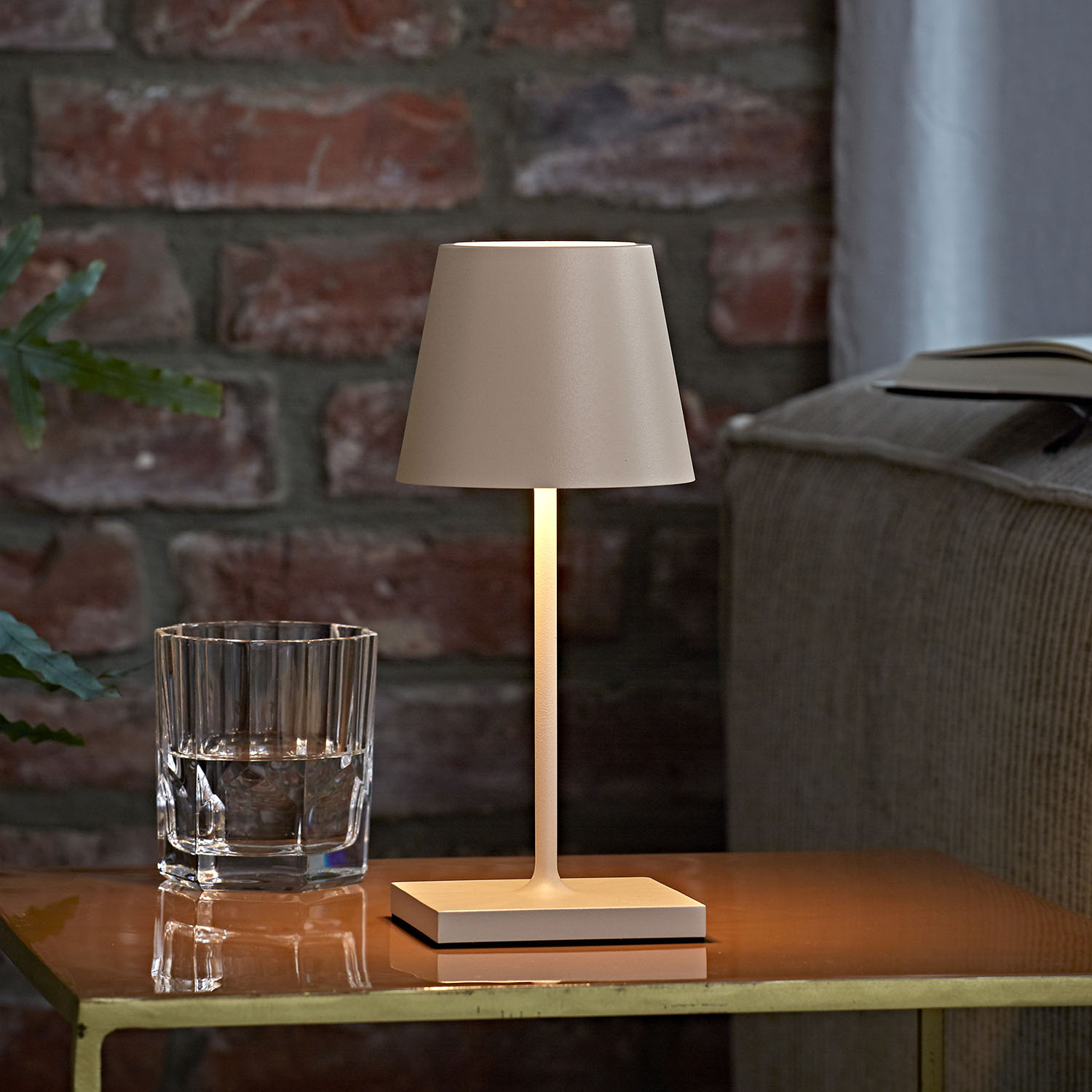 Eine beleuchtete Lampe auf einem Tisch neben einem Glas Wasser und einem Buch, mit Ziegelwand im Hintergrund.