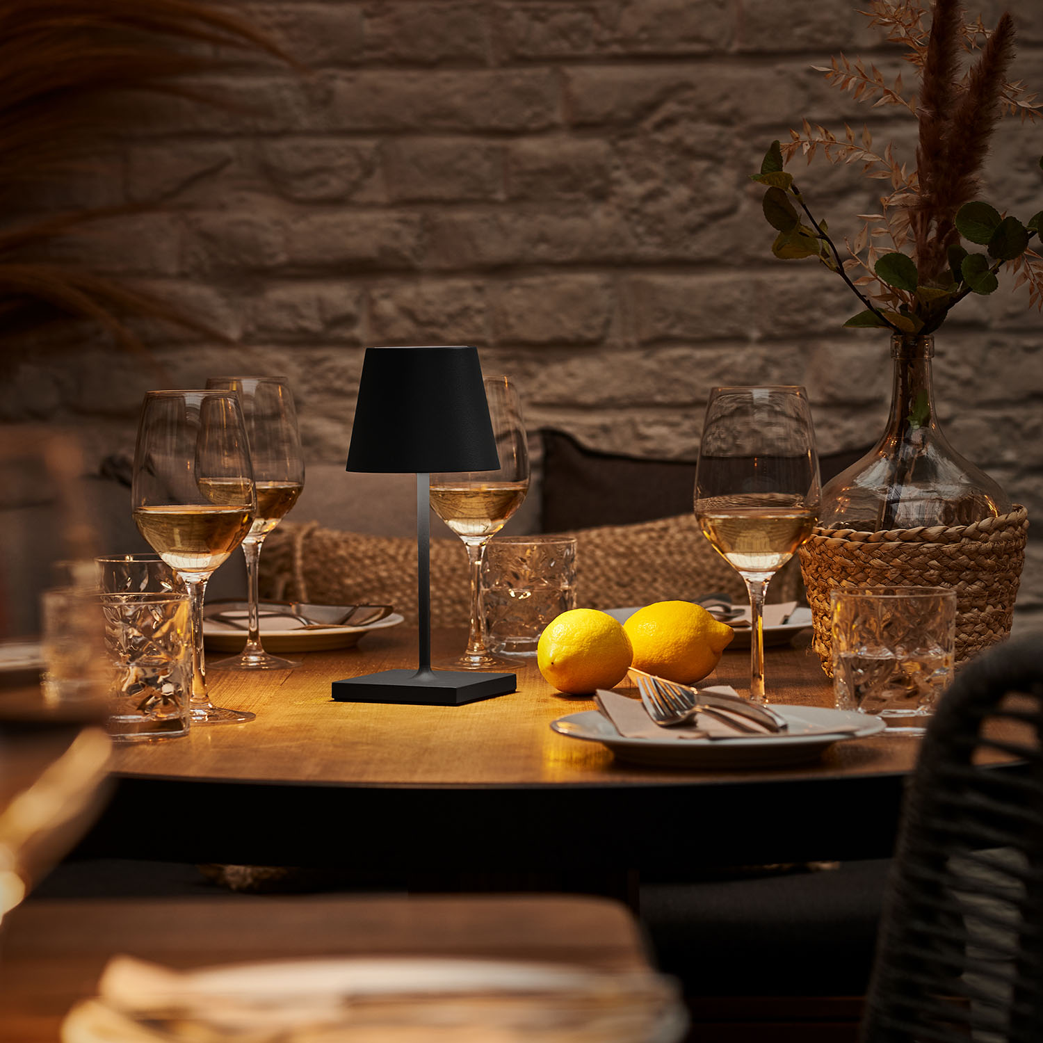 Tisch mit Lampe, Gläsern, Zitronen und Vase vor einer Backsteinmauer.