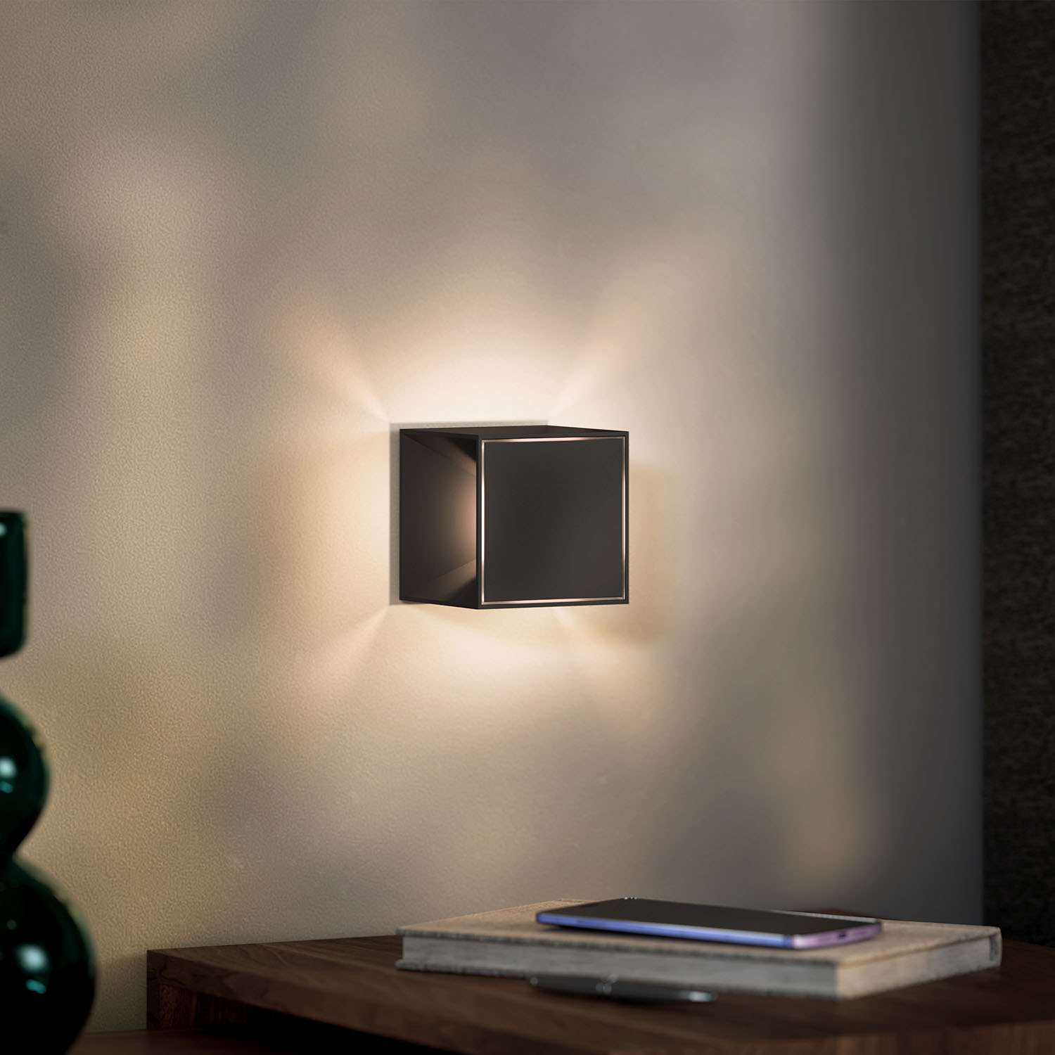 Eine schwarze quadratische Wandleuchte an einer weißen Wand. Licht scheint um die Lampe herum.