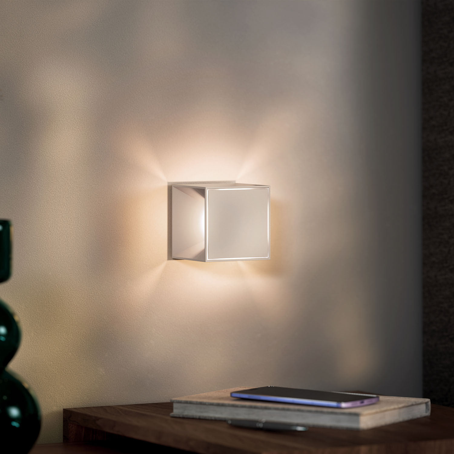 Eine quadratische Lampe an einer Wand. Ein Telefon und Buch auf einem Tisch.