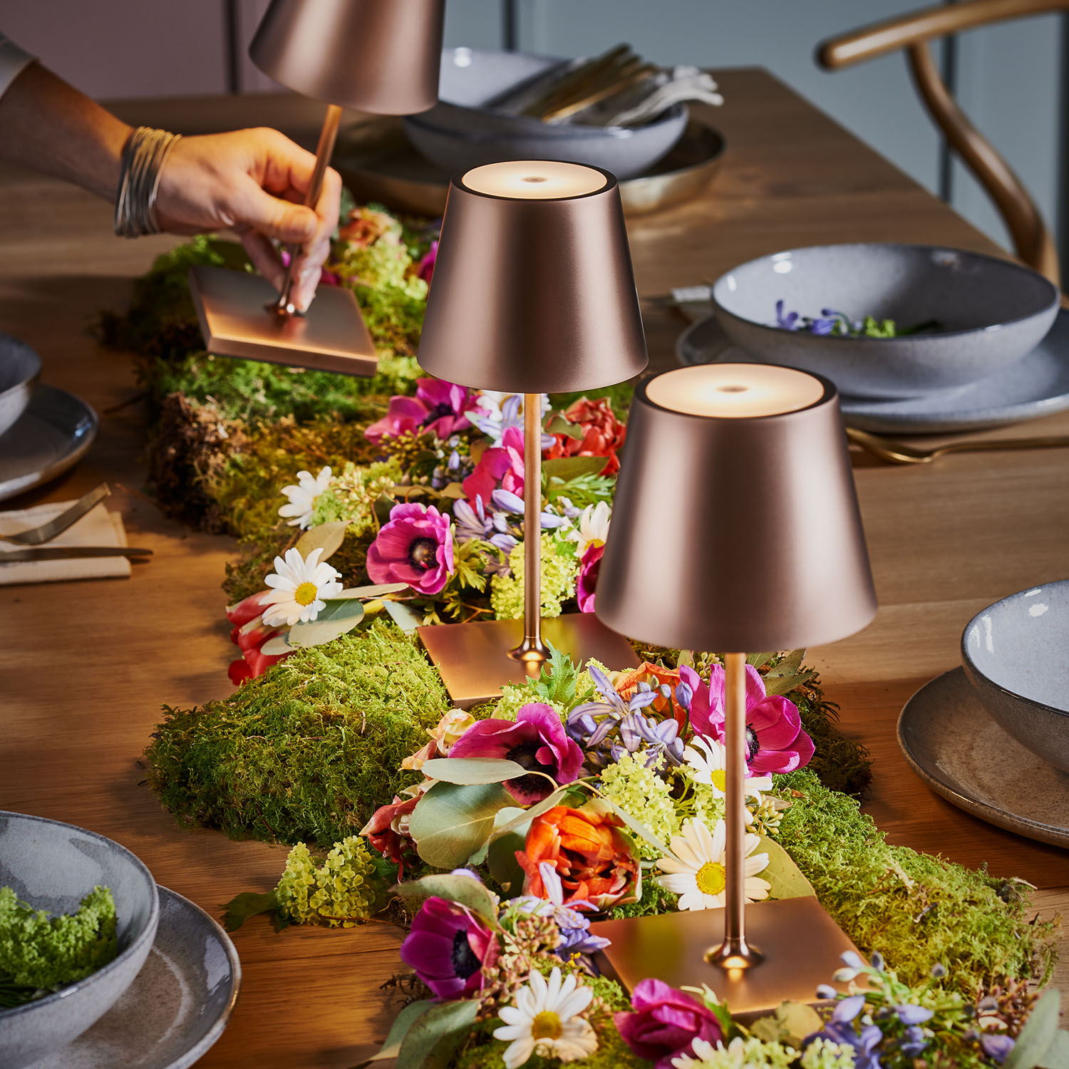 Zwei Kupferlampen auf einem Tisch mit Blumen und Moos. Eine Hand schreibt auf eine Kupferplatte.
