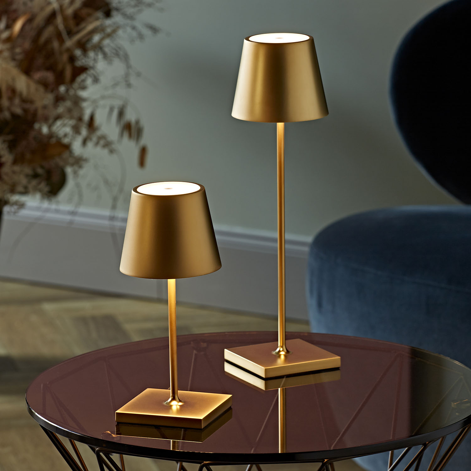 Zwei goldfarbene Lampen mit quadratischen Sockeln auf einer reflektierenden Oberfläche, mit einem blauen Stuhl im Hintergrund.