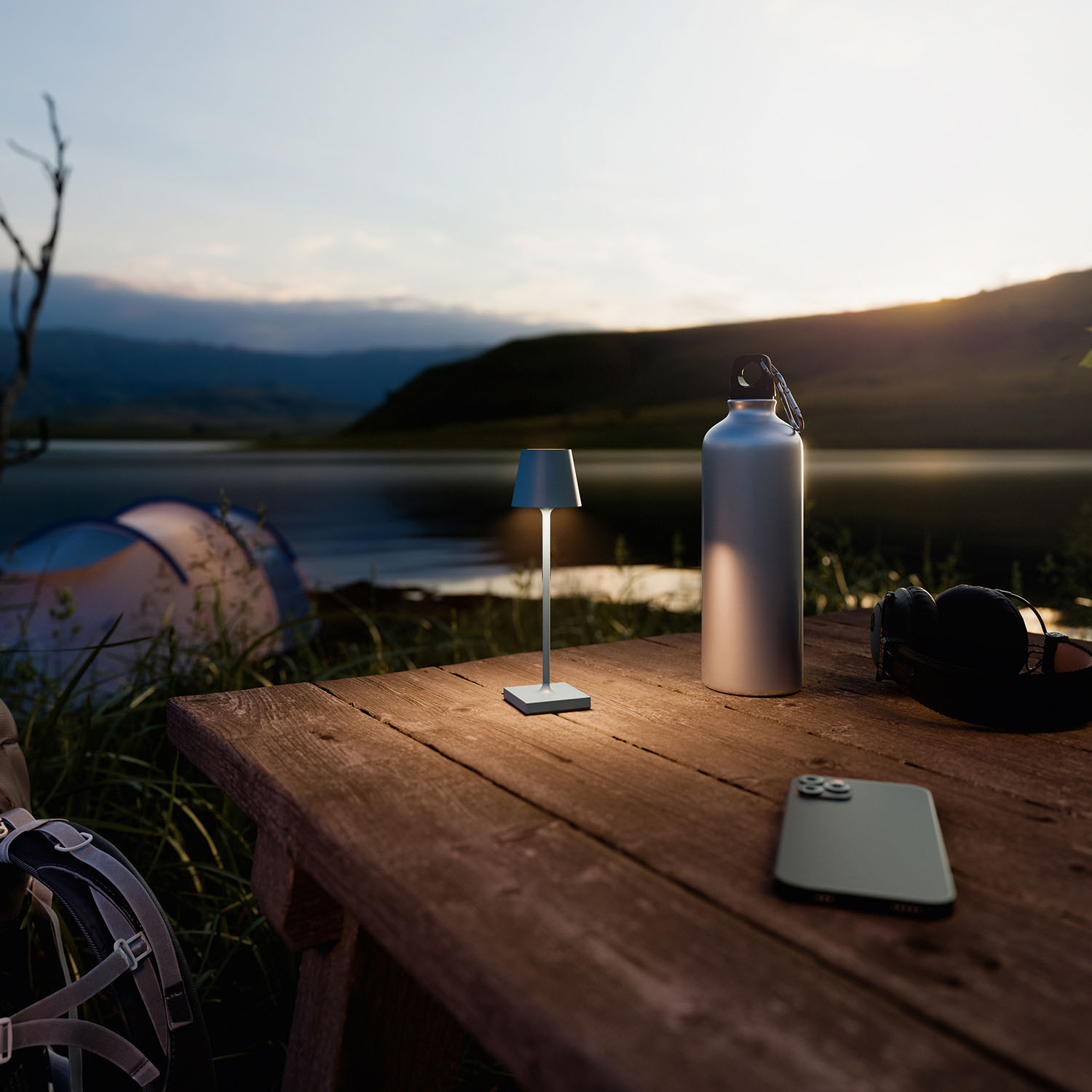 Ein Tisch mit Lampe, Wasserflasche, Telefon und Kopfhörern steht neben einem See bei Sonnenuntergang.