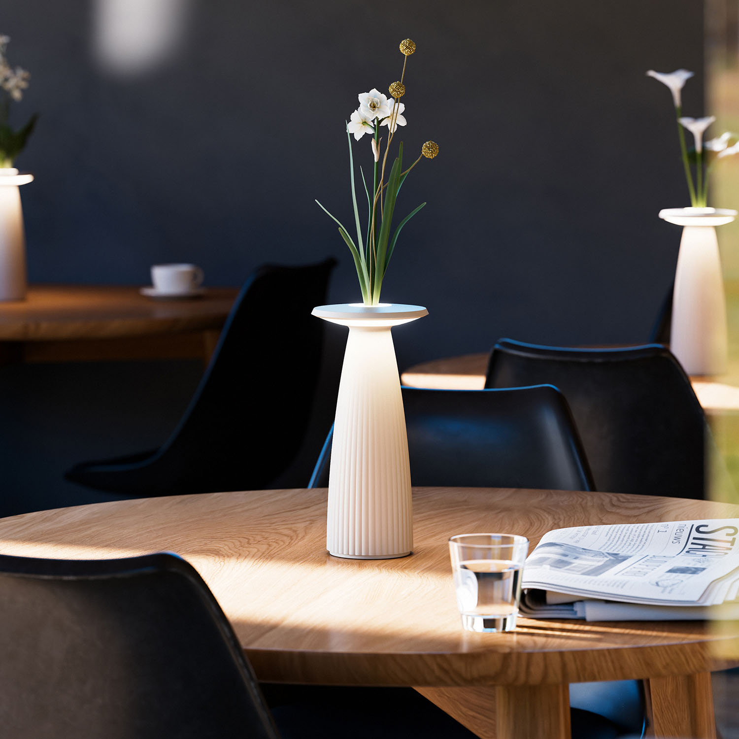 Weiße Lampe auf Holztisch, Vase mit Blumen, Glas Wasser, Zeitung und schwarze Stühle im Hintergrund.