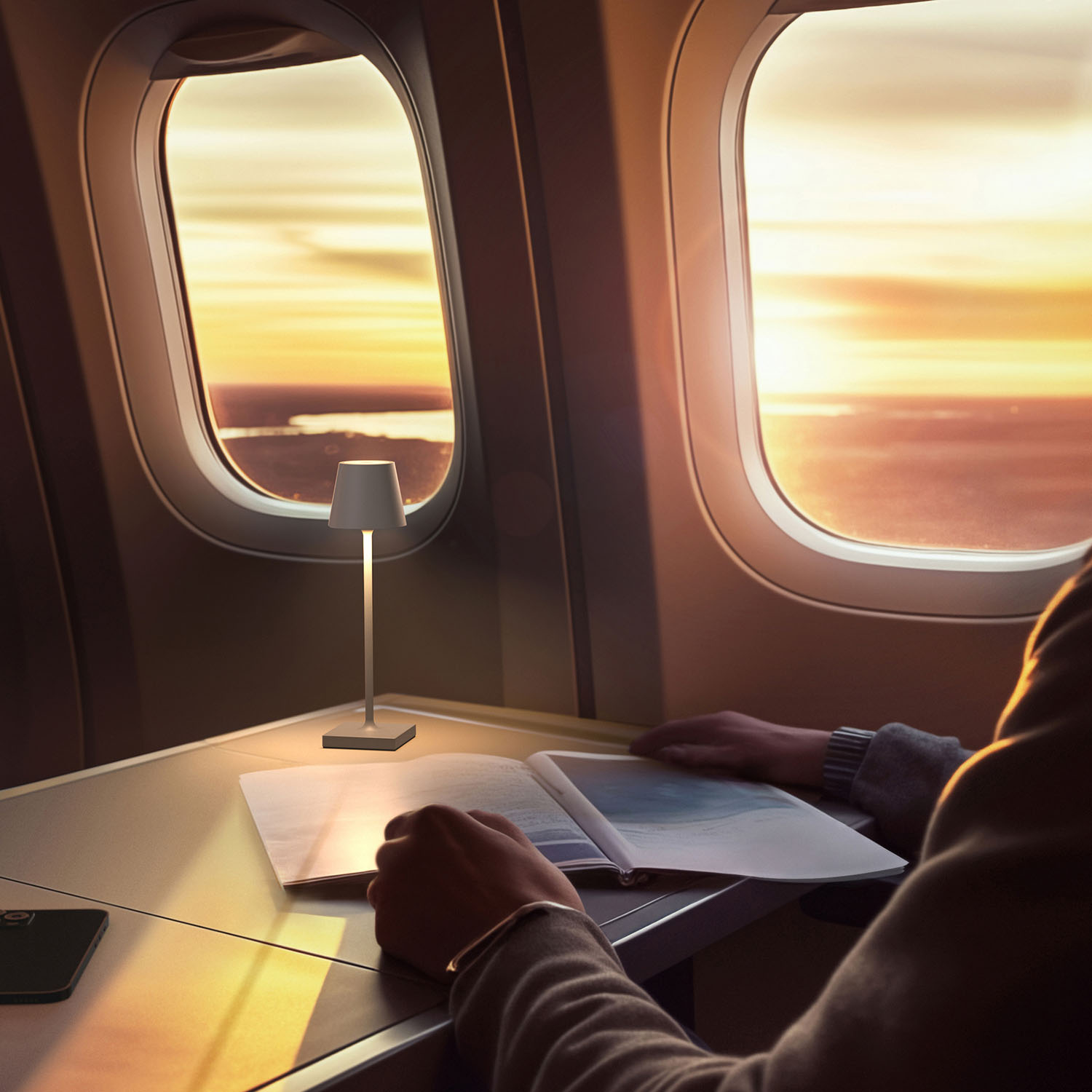 Ein Tisch mit Lampe, Buch und Telefon, neben einem Flugzeugfenster mit Sonnenuntergang.