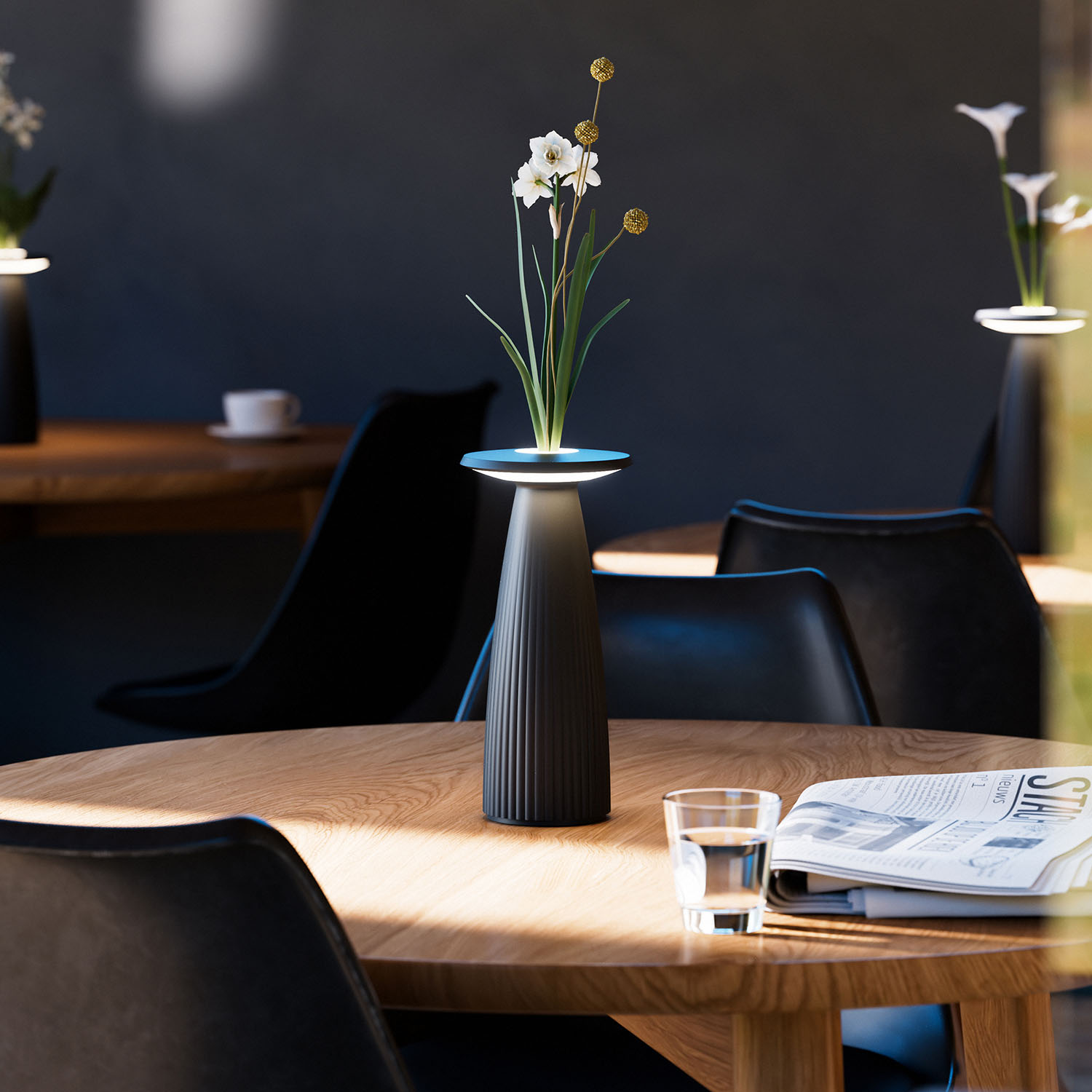 Eine schwarze Vase mit Blumen auf einem Holztisch. Gläser und eine Zeitung sind vorhanden.