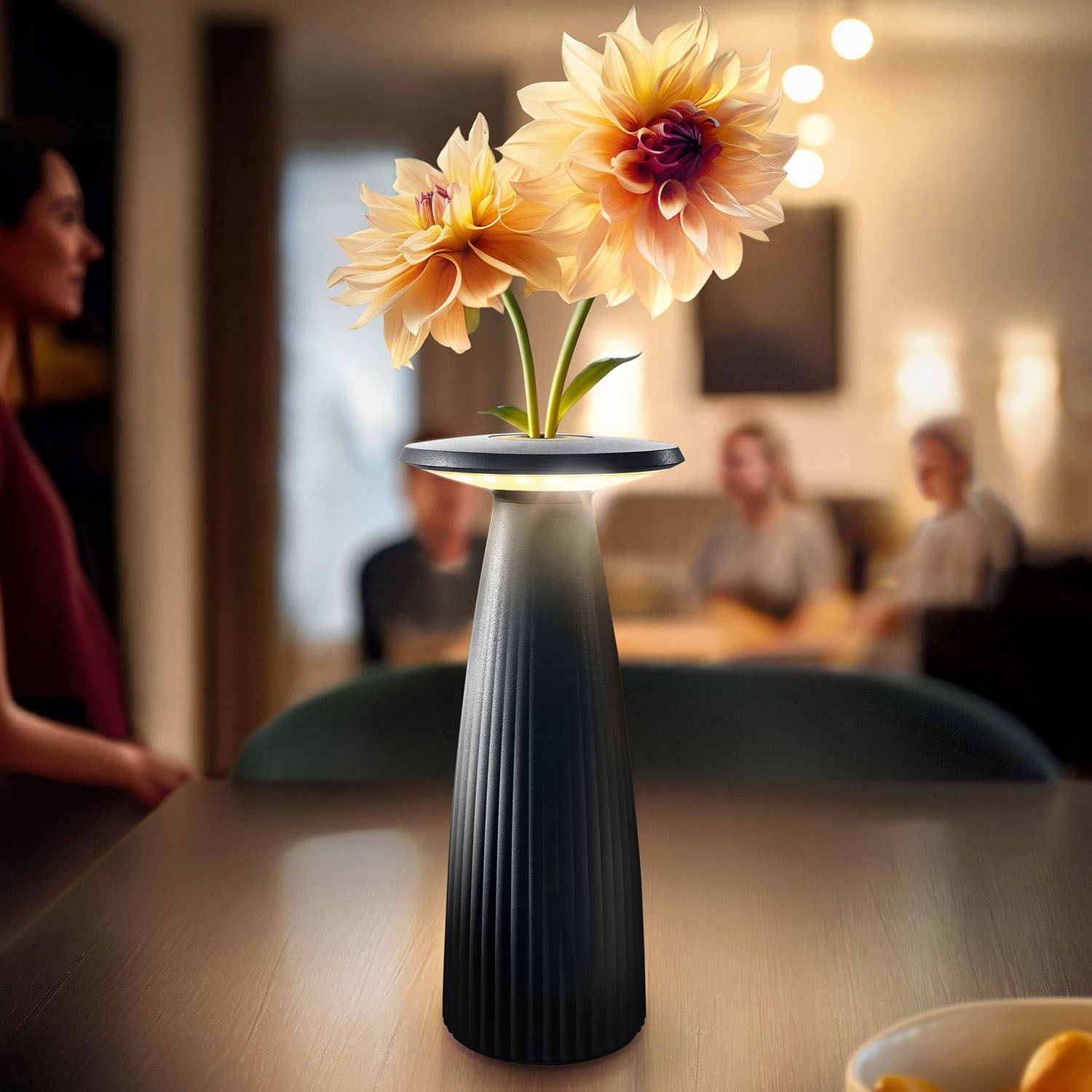 Eine hohe schwarze Vase mit beleuchteten Blumen auf einem Tisch, mit Personen im Hintergrund, weichgezeichnet.