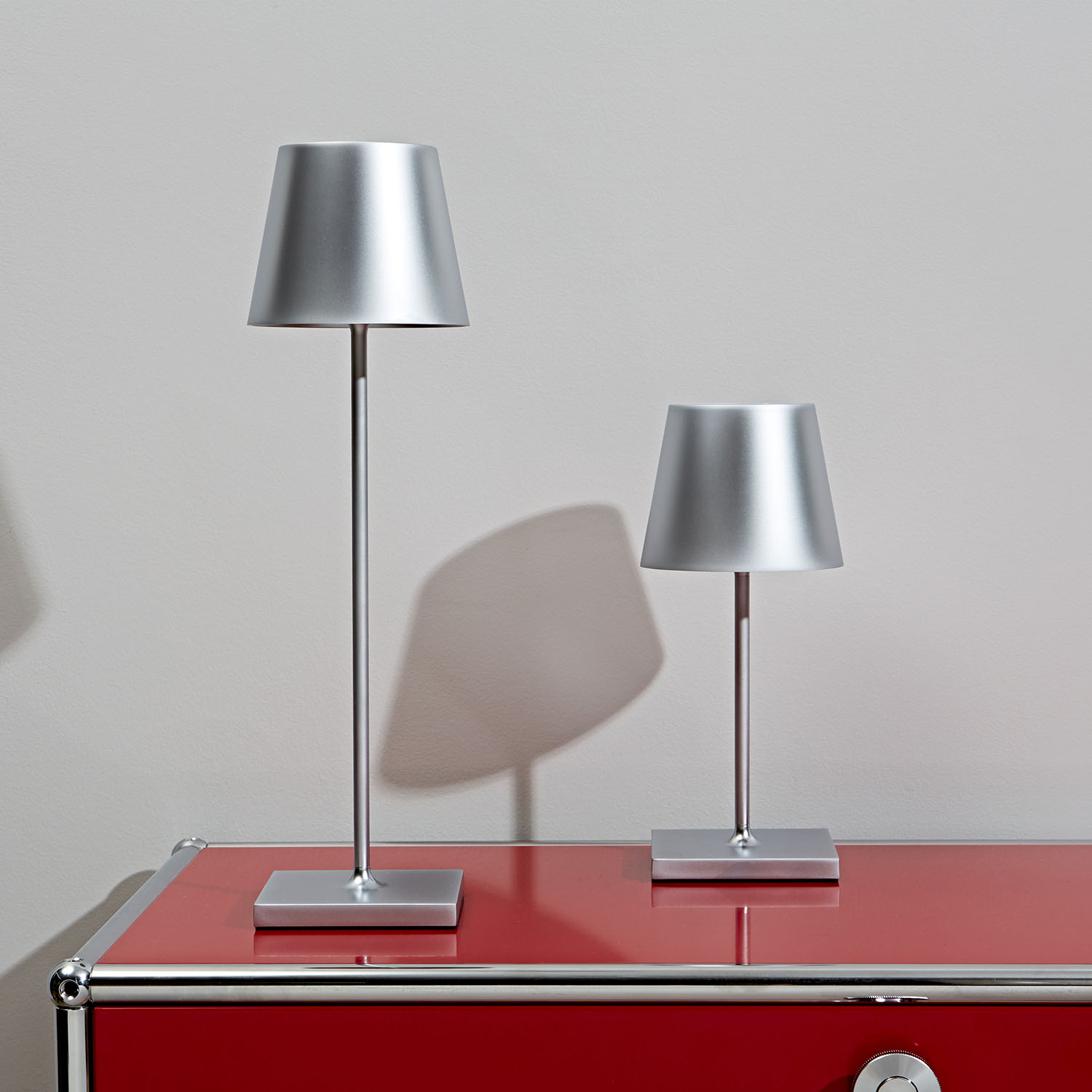 Zwei silberne Lampen, eine hohe und eine kurze, auf einem roten Schrank vor einer grauen Wand.