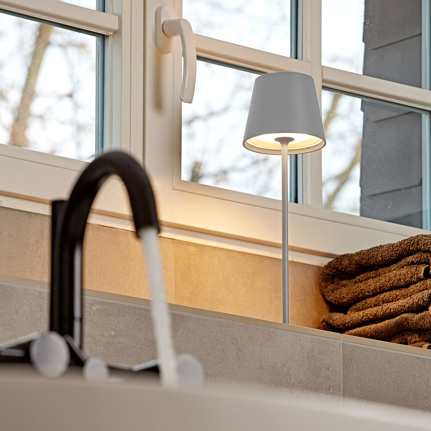 Eine Lampe am Fenster. Die Lampe ist grau und leuchtet, ein Wasserhahn und Handtücher sind sichtbar.