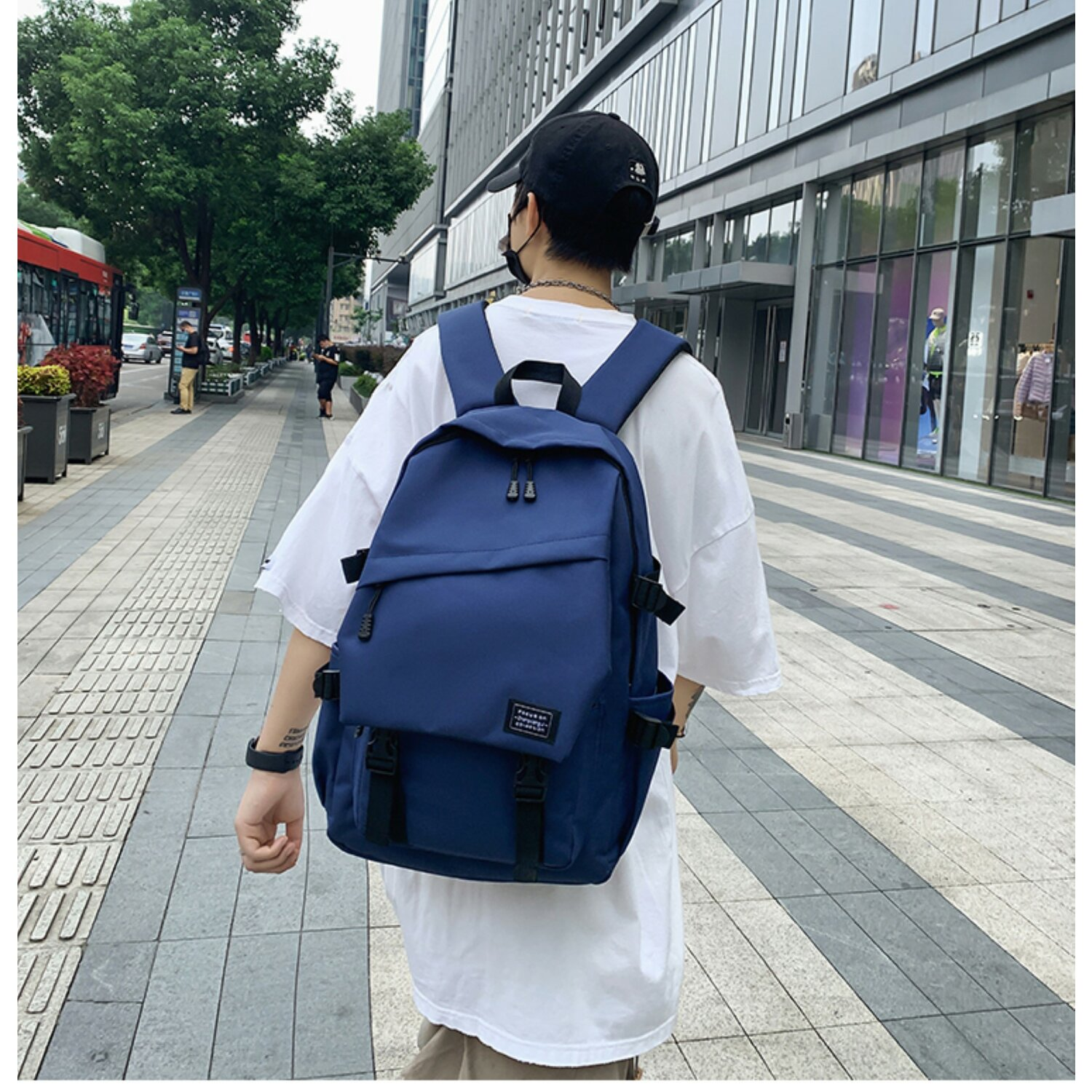 Ein Mann mit Rucksack geht vor einem Stadtgebäude, trägt Maske und weißes T-Shirt.