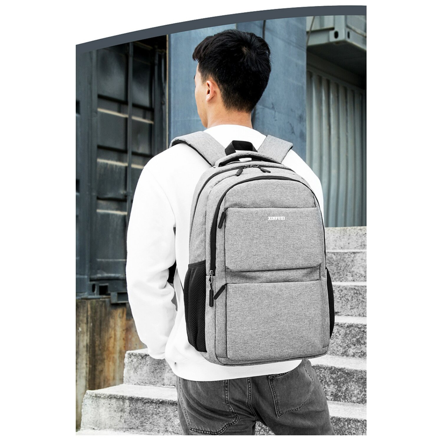 Ein Mann mit grauem Rucksack, steht vor einem grauen Gebäude. Rückansicht.