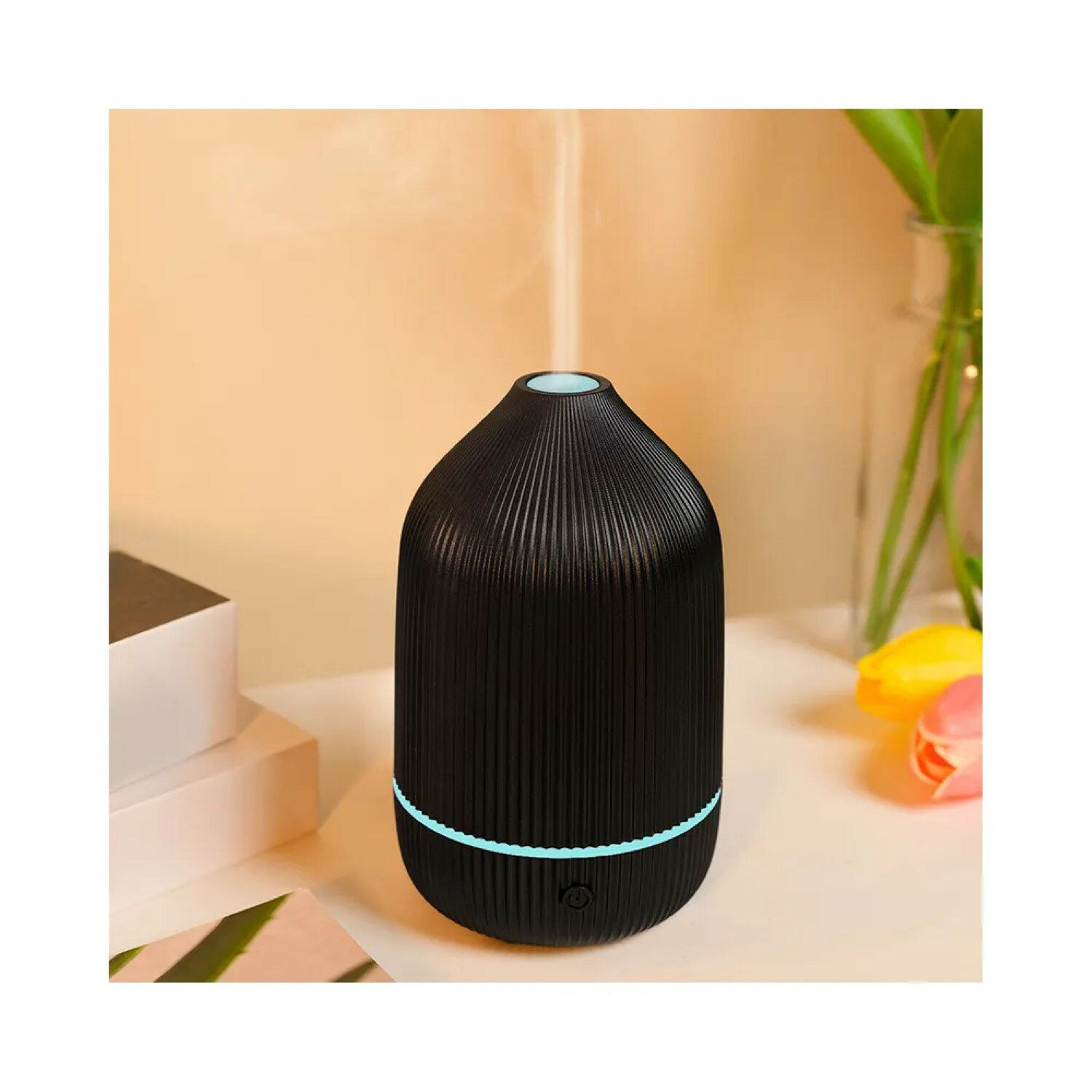 Ein schwarzer Diffusor mit Dampf, auf einem weißen Tisch. Im Hintergrund Blumen.