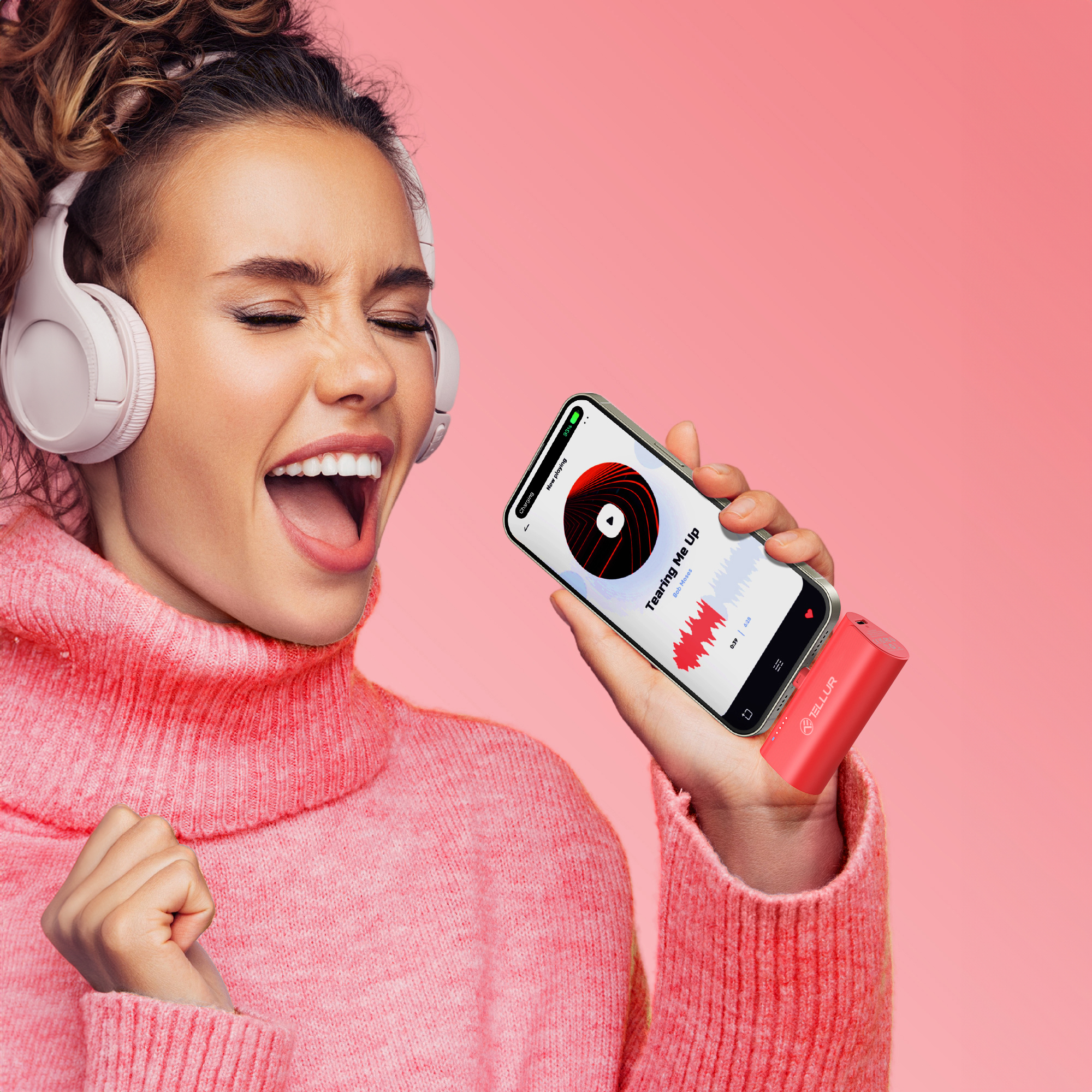 Kobieta śpiewa, trzymając telefon z aplikacją muzyczną. Różowe tło. Białe słuchawki i czerwony odtwarzacz muzyki.