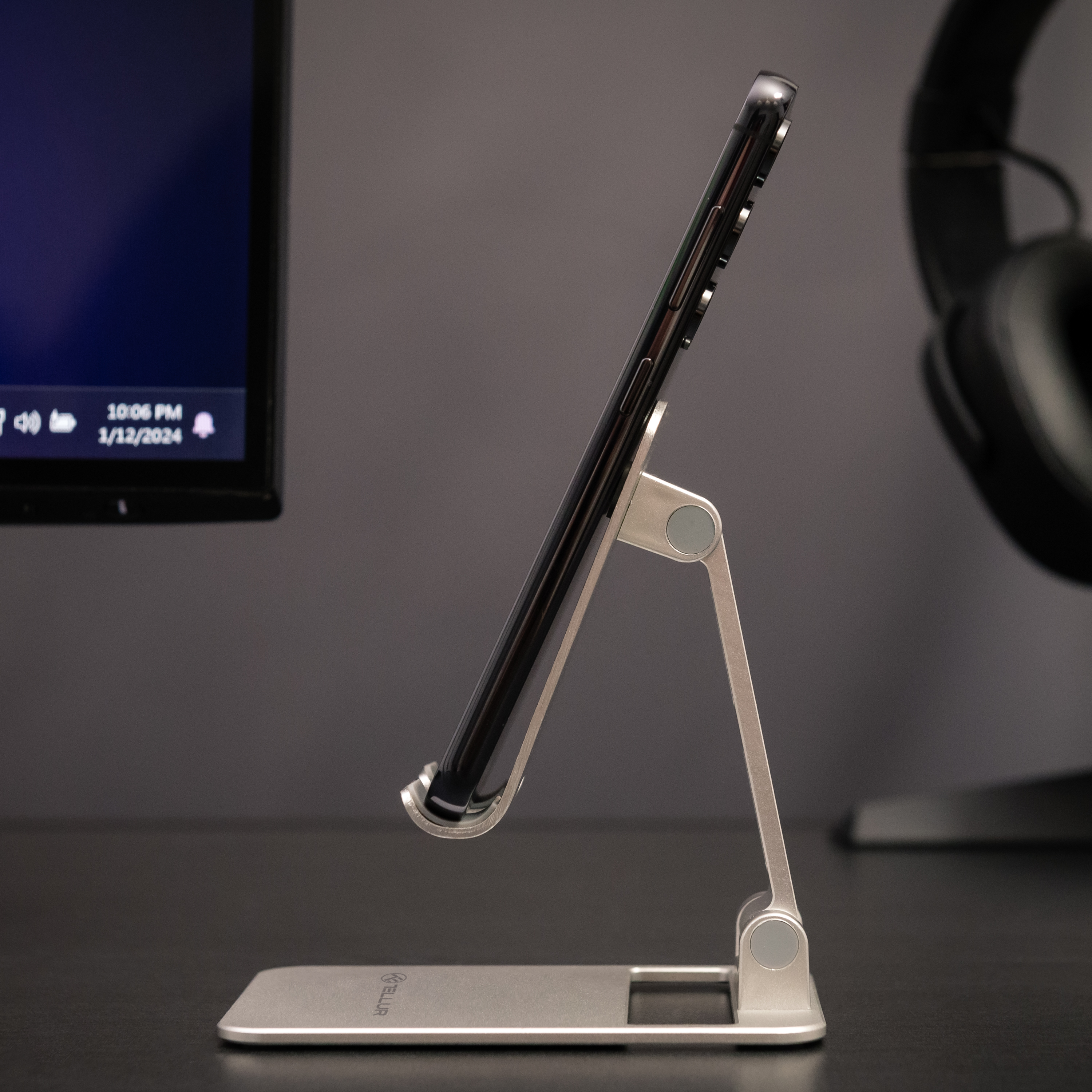 Srebrny stojak na telefon trzymający czarny telefon. Ekran widoczny. Ciemne tło biurka.