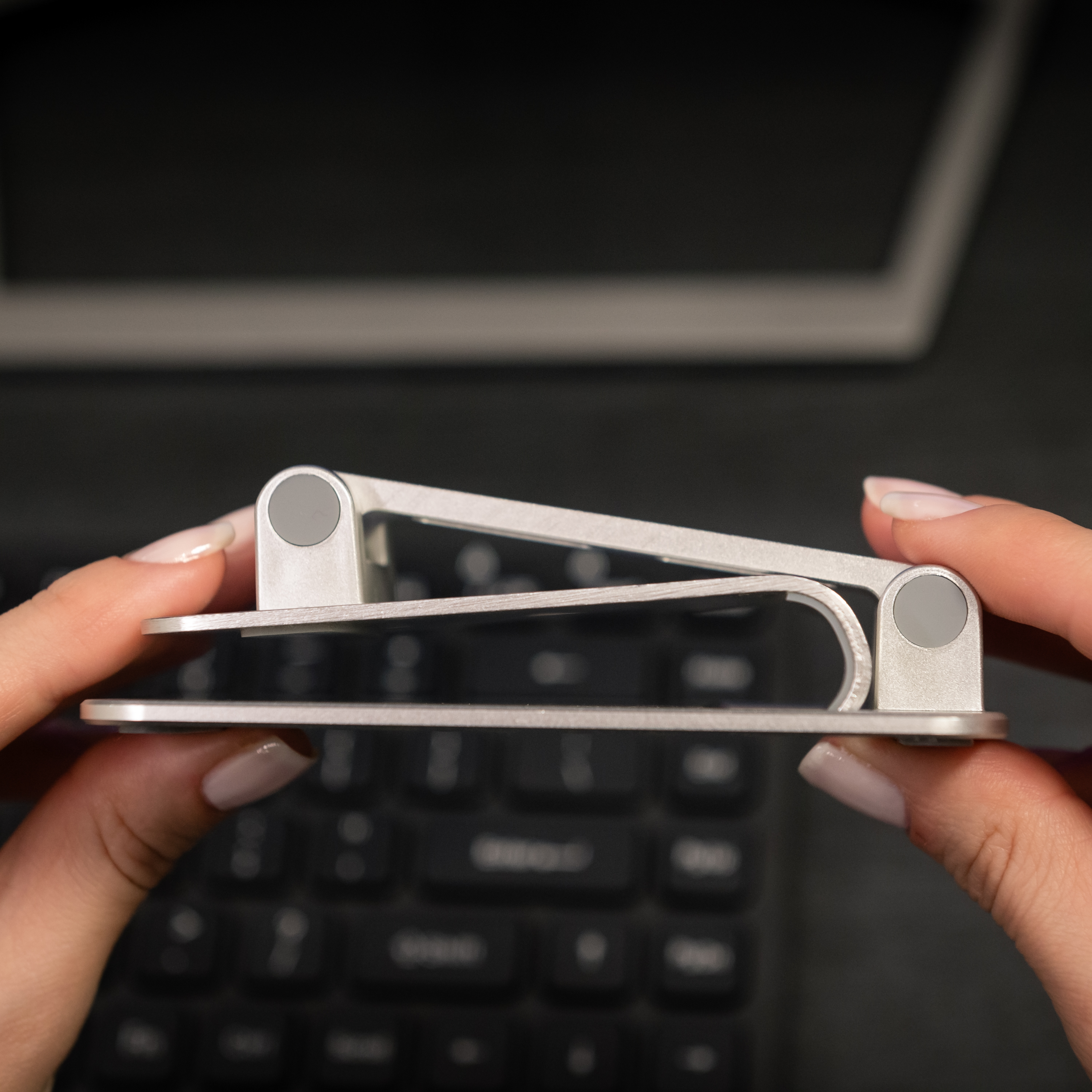 Dłonie trzymające srebrną podstawkę pod laptopa nad klawiaturą. Szare tło.