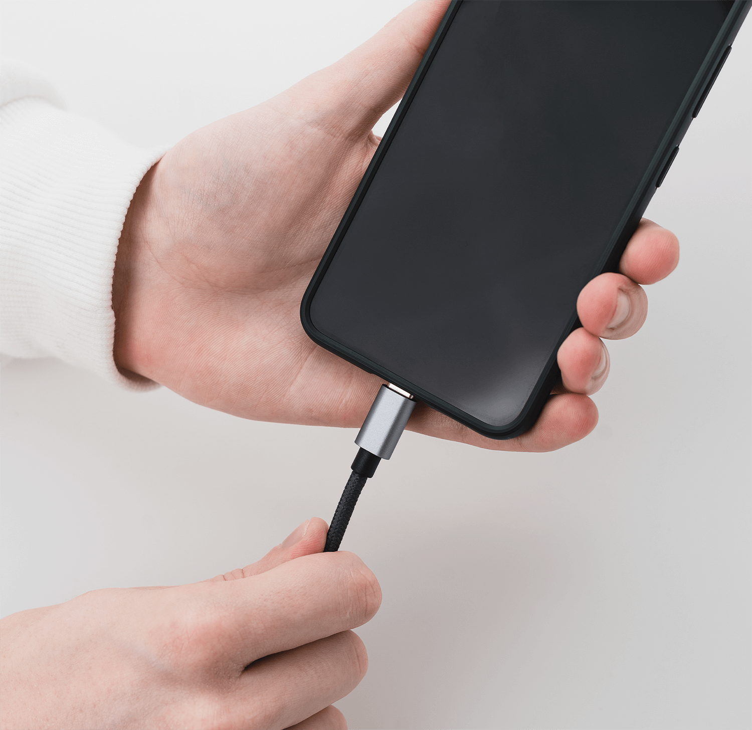Ręce osoby trzymającej czarny telefon z podłączonym kablem ładującym. Białe tło.