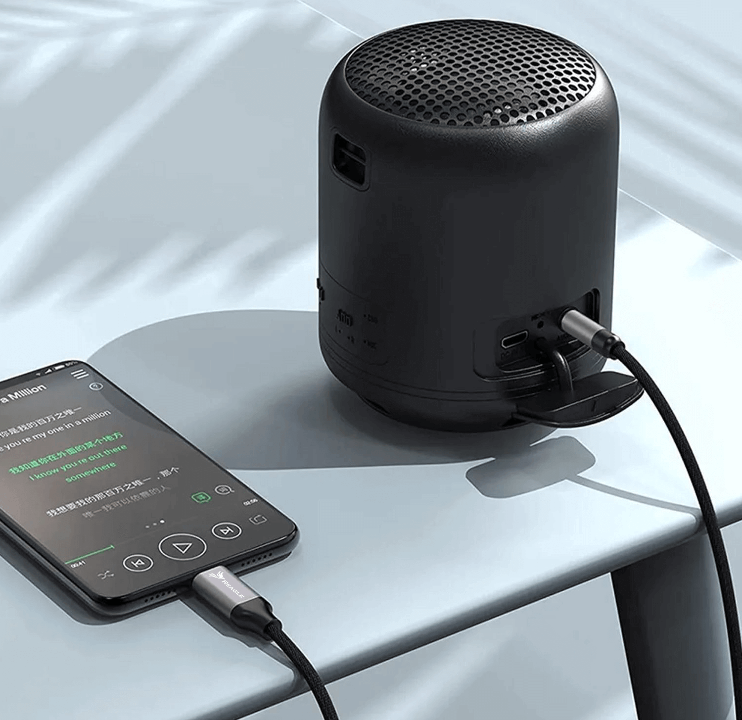 Czarny głośnik podłączony do smartfona kablem na jasnoniebieskiej powierzchni.