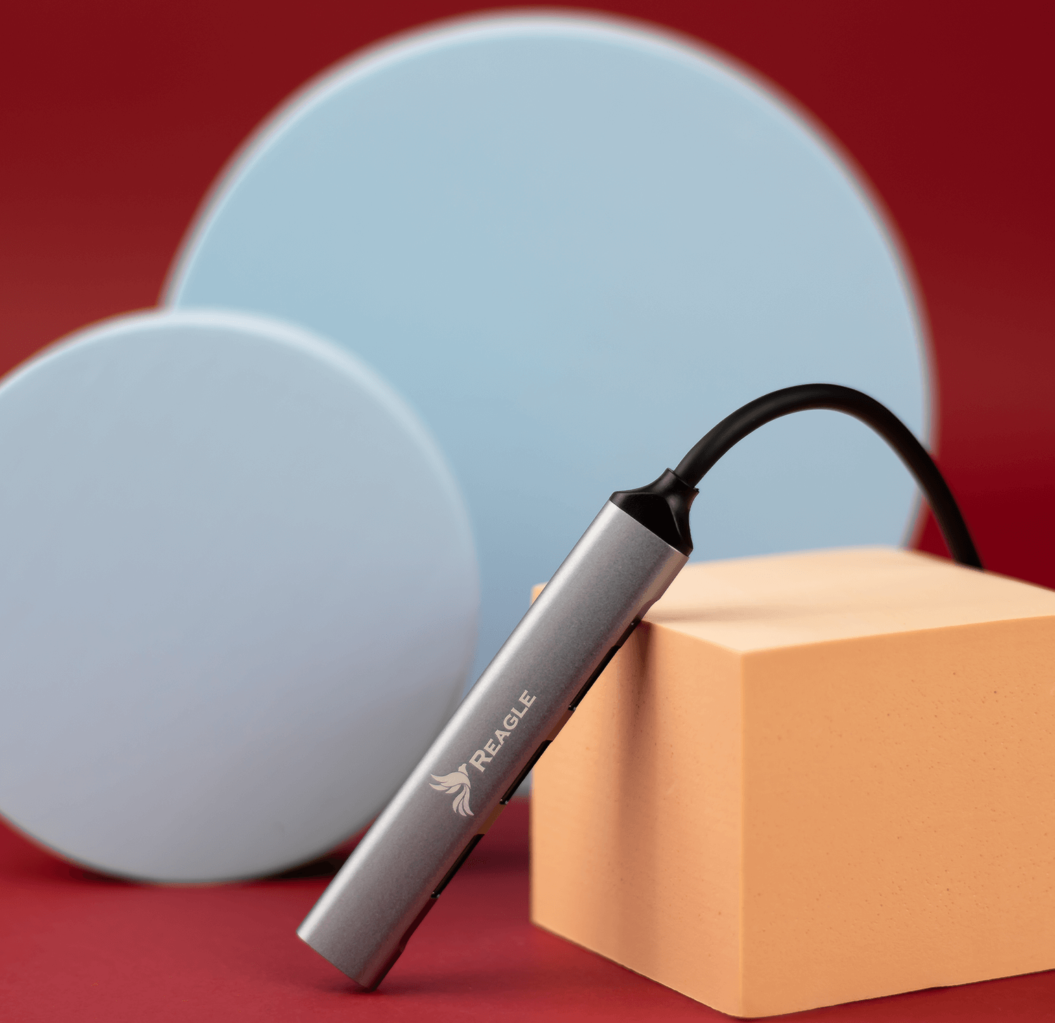 Srebrny adapter leży na czerwonej powierzchni, obok niebieskich i pomarańczowych kształtów. Wychodzi kabel.