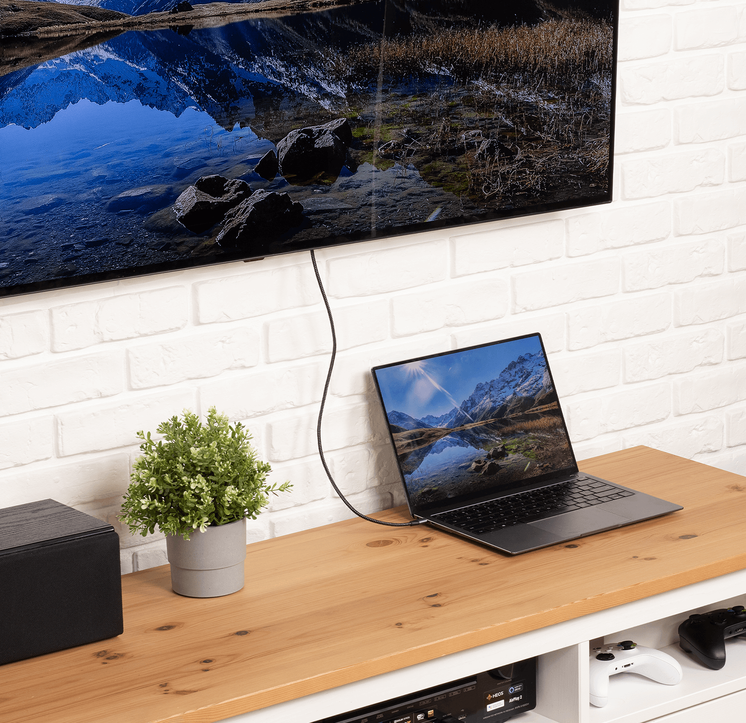 Telewizor, laptop i roślina stoją na drewnianym stole. Za nimi biała ceglana ściana.