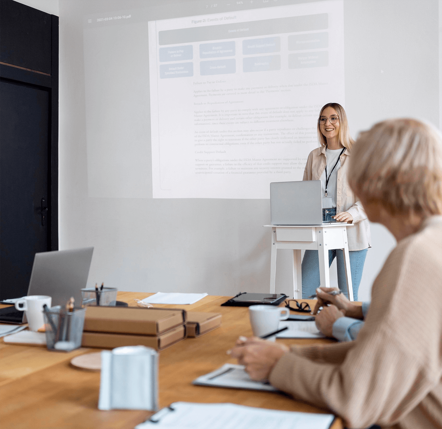 Kobieta prezentuje na spotkaniu. Stoi przy stole z laptopem, twarzą do publiczności.