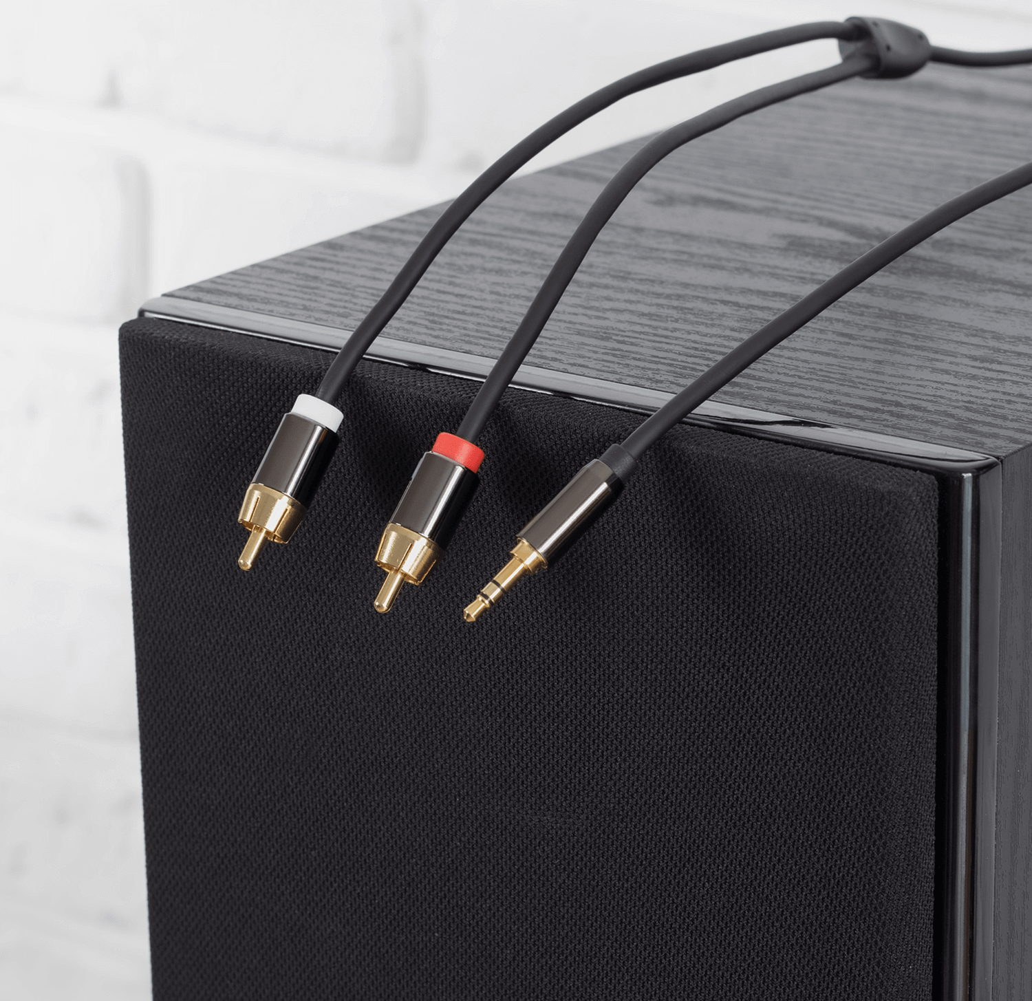 Głośnik z trzema podłączonymi kablami, czarno-złote złącza, na białej ceglanej ścianie.