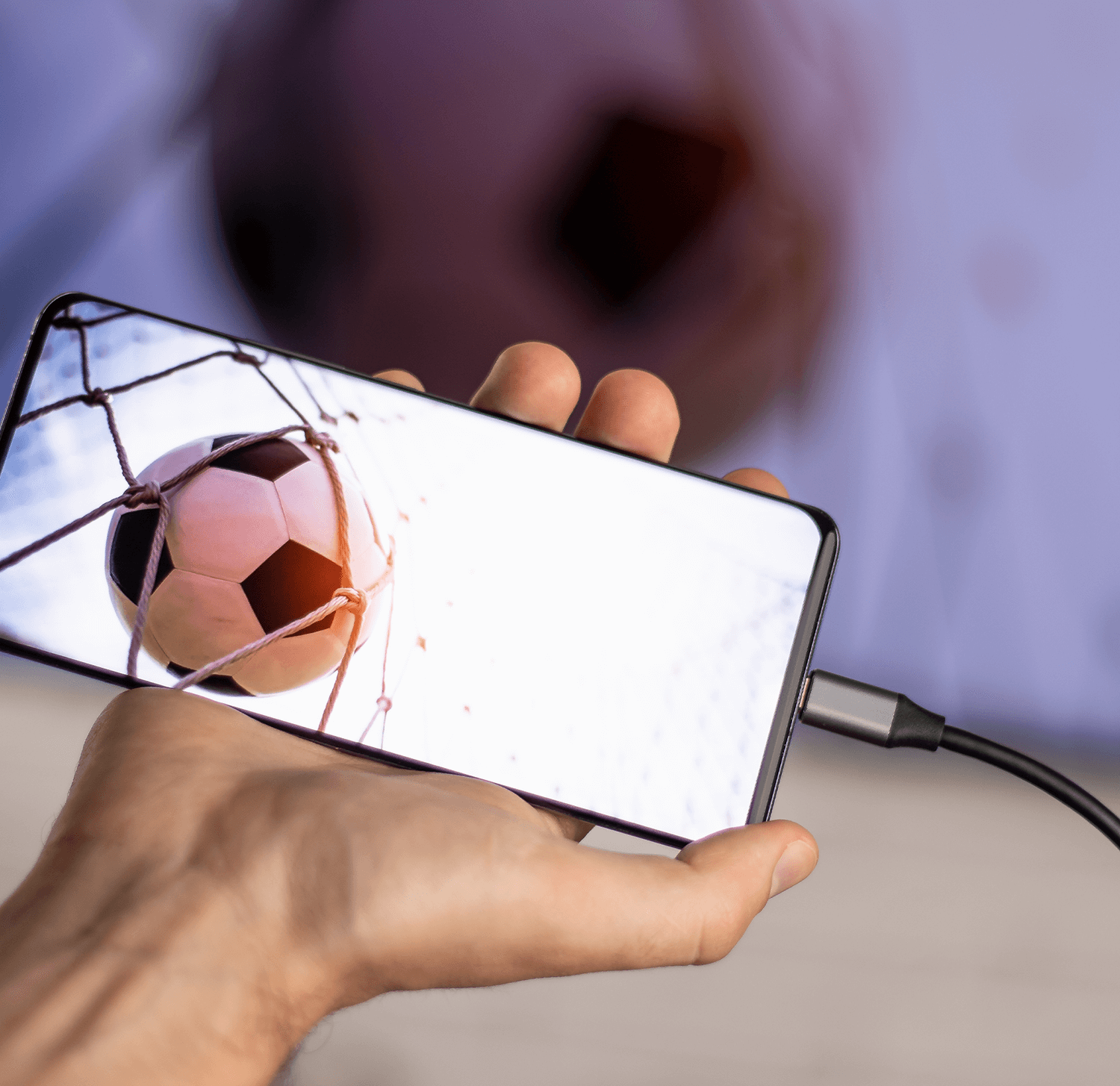 Ręka trzymająca smartfon. Mecz piłki nożnej na ekranie. Podłączony czarny kabel.