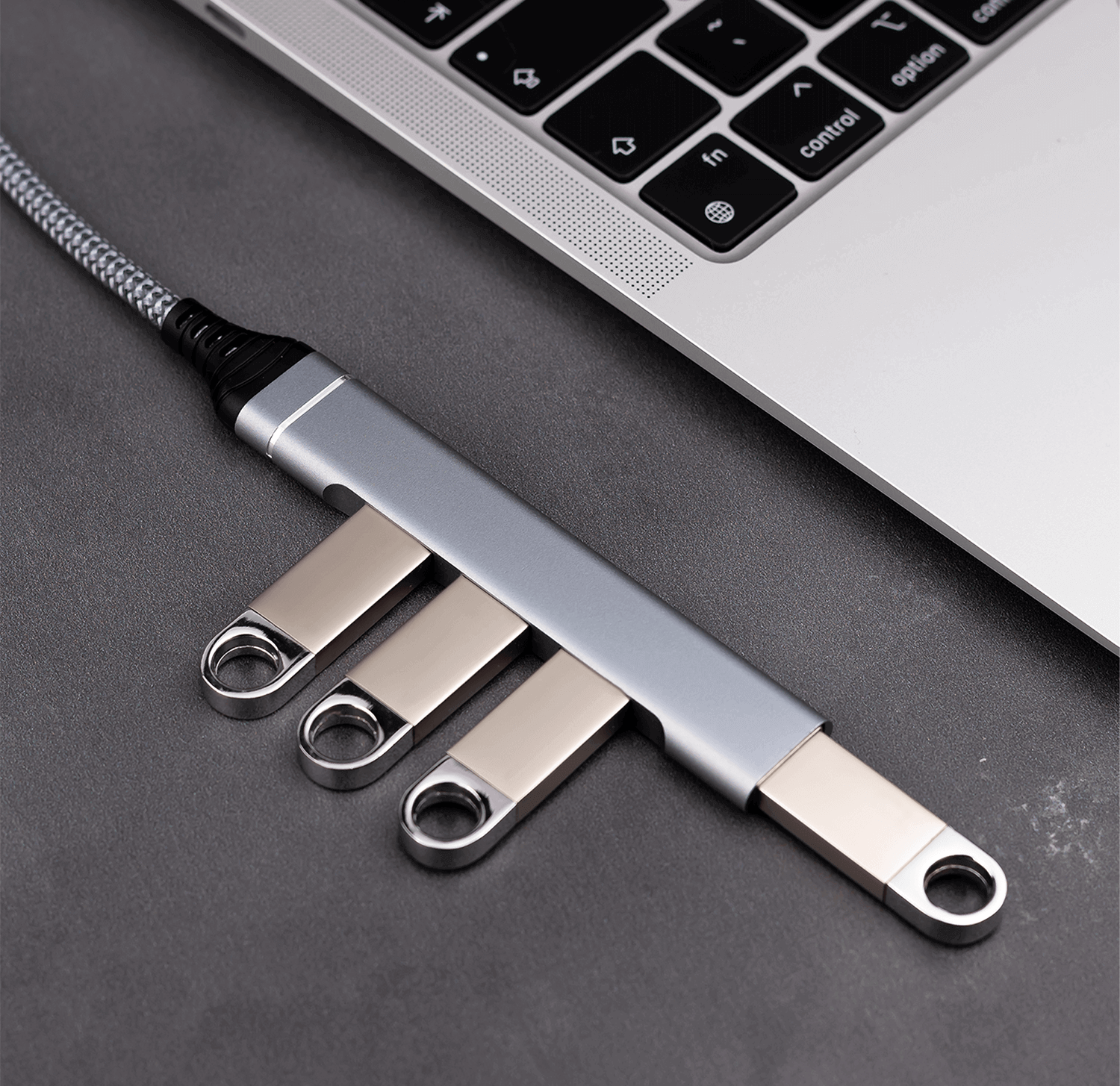 Laptop z srebrnym hubem USB-C i trzema pendrive'ami na szarej powierzchni.