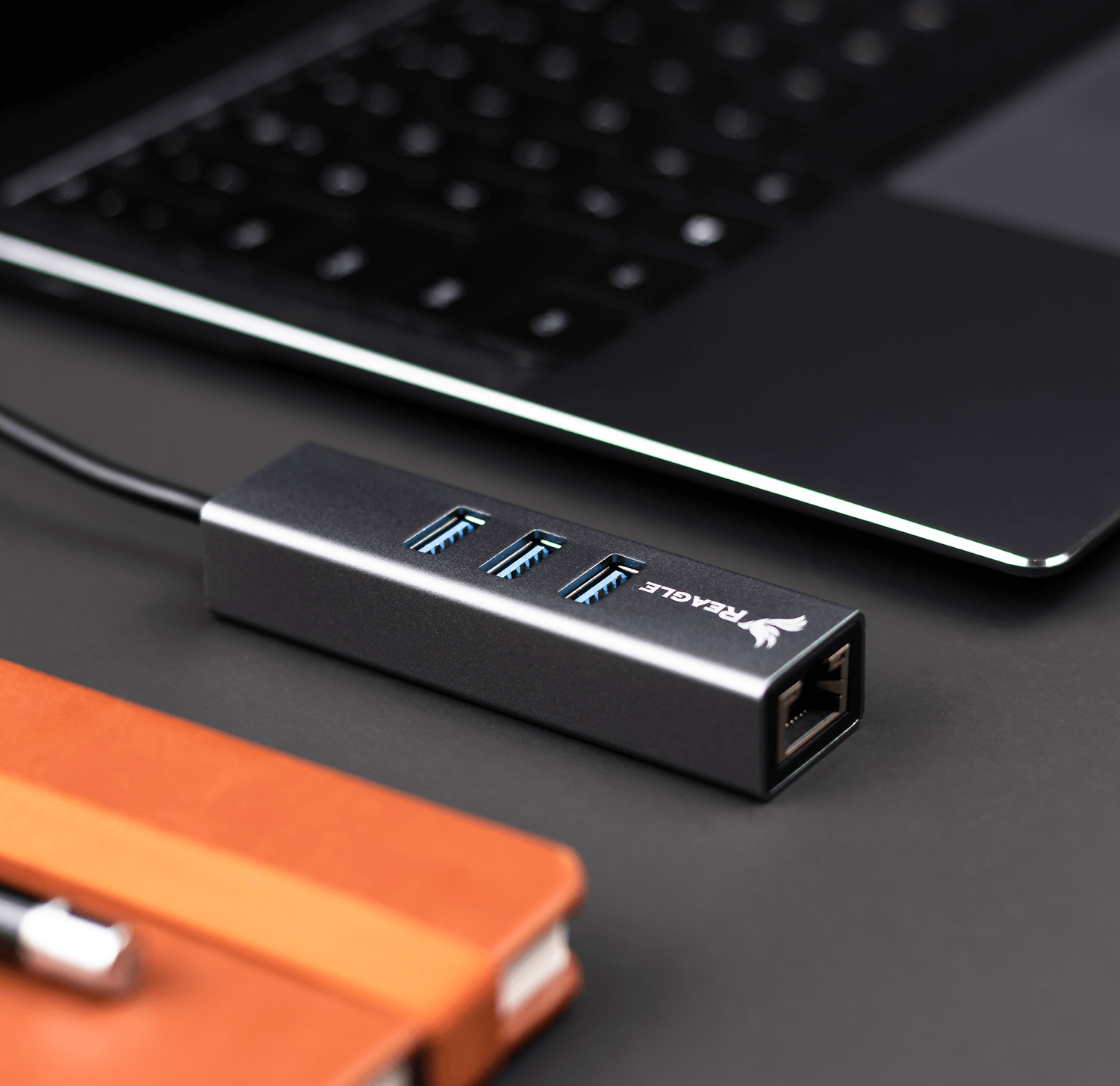 Szary hub USB Reagle z trzema portami, laptopem i notatnikiem na ciemnej powierzchni.