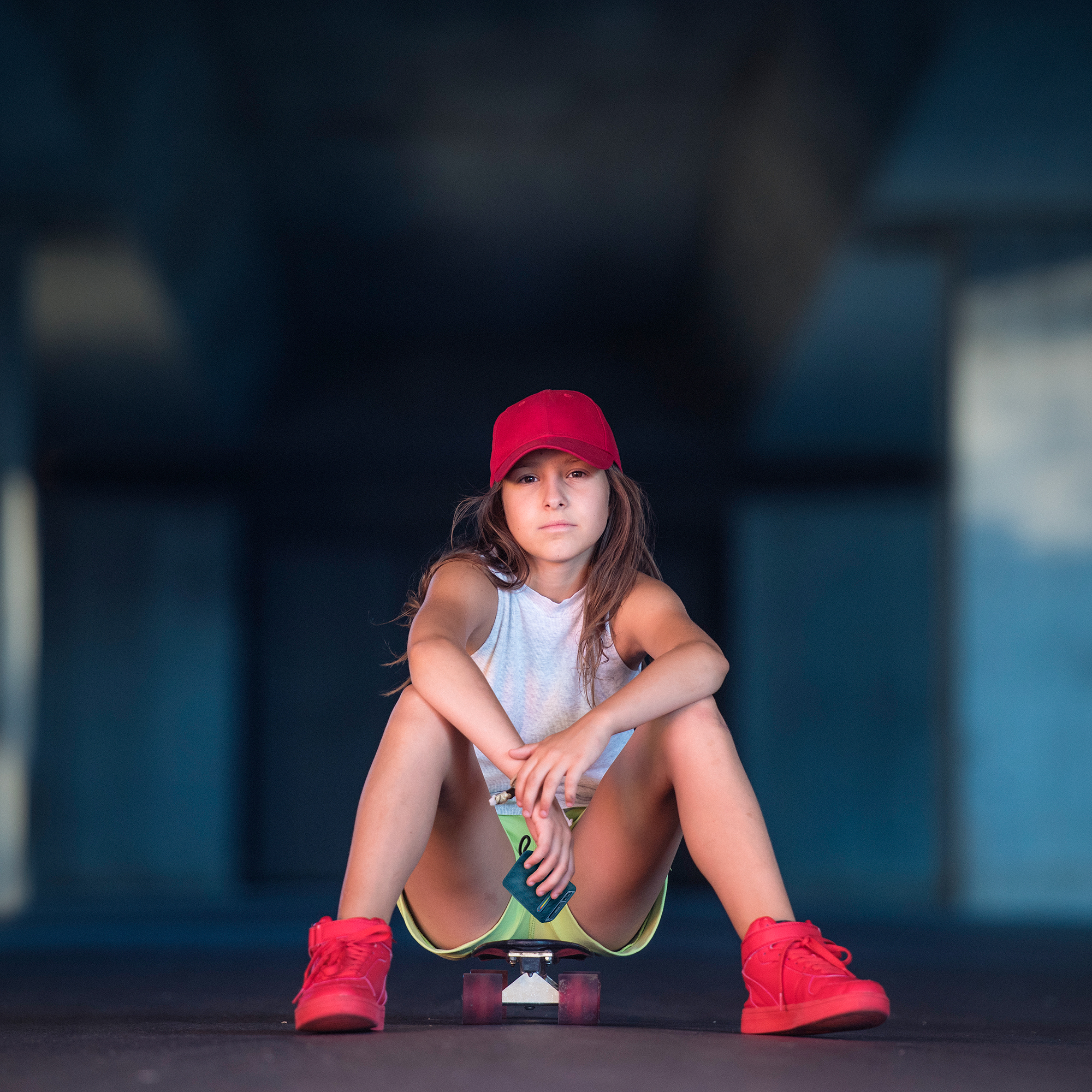 Dziewczyna w czerwonej czapce siedzi na deskorolce pod mostem, trzymając butelkę.