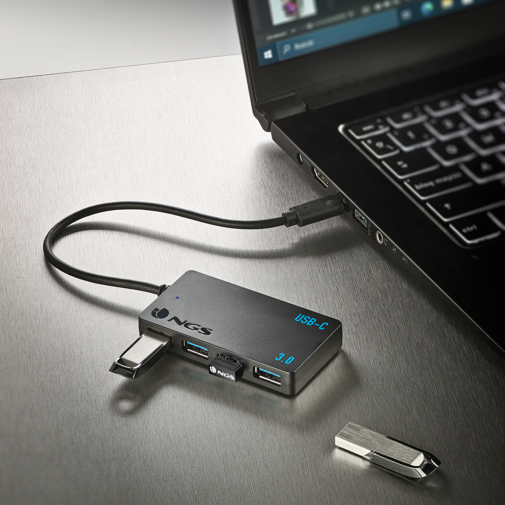 Ein USB-C-Hub und ein USB-Laufwerk sind auf einer Metalloberfläche mit einem Laptop verbunden.