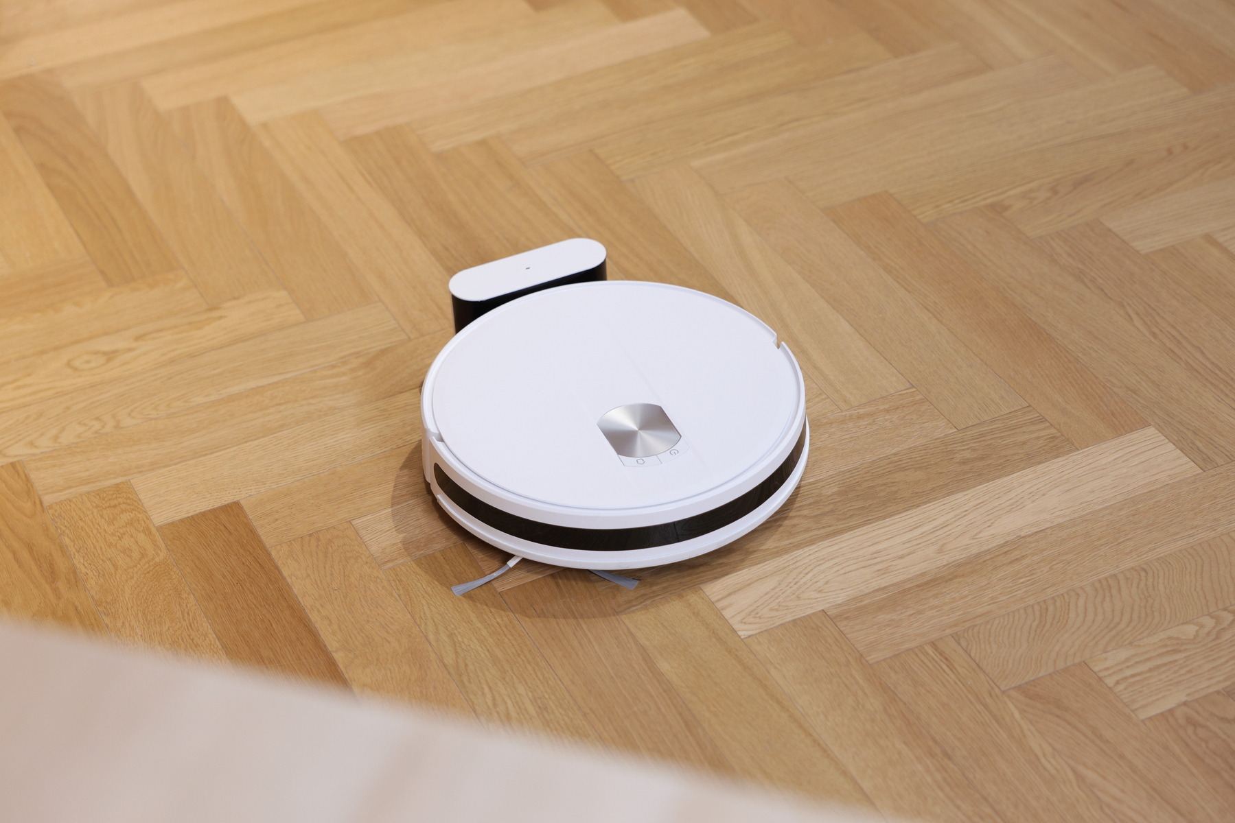 Biały odkurzacz robot na drewnianej podłodze, ze stacją ładującą w tle.