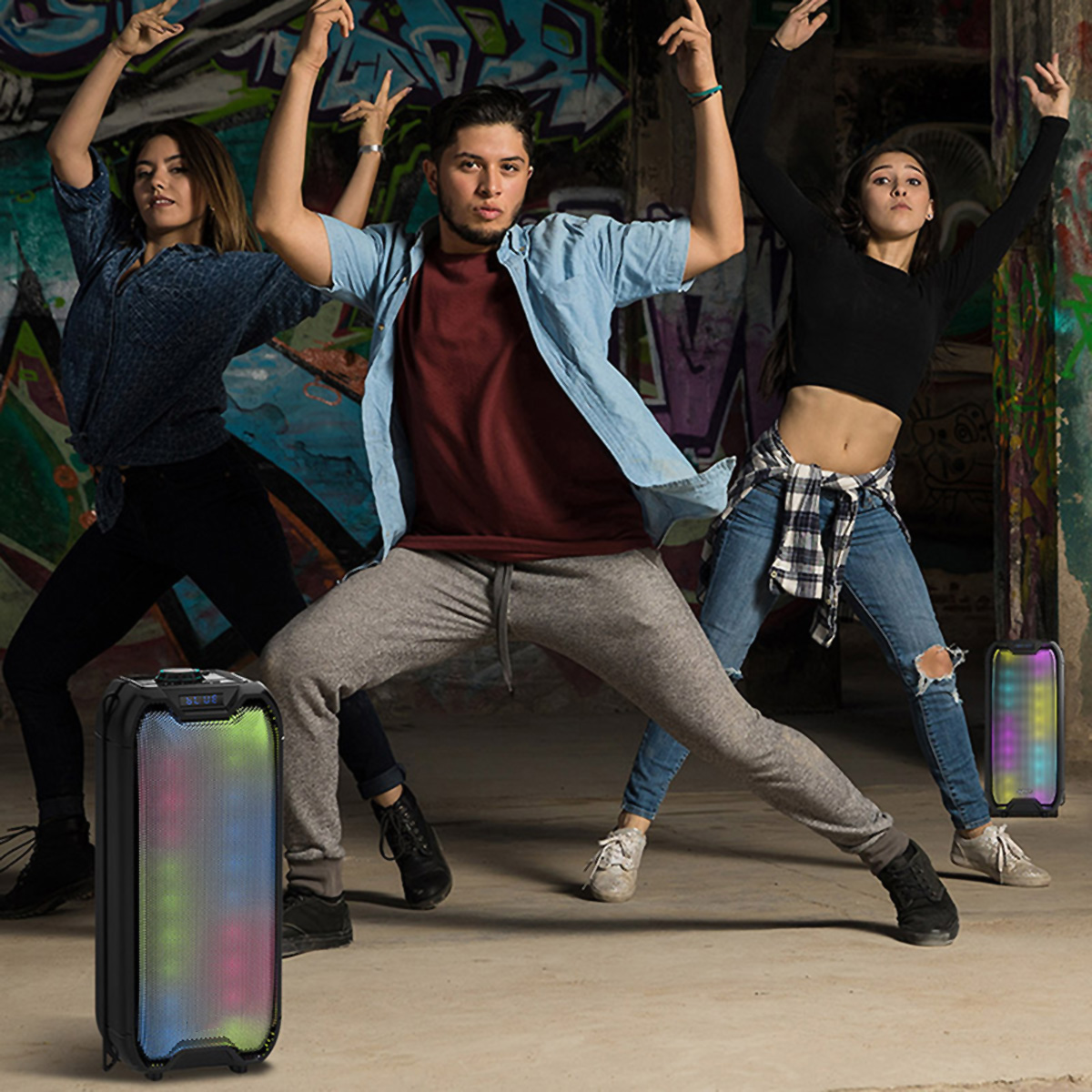 Tres personas bailando con los brazos levantados, dos altavoces en primer plano, fondo de graffiti.