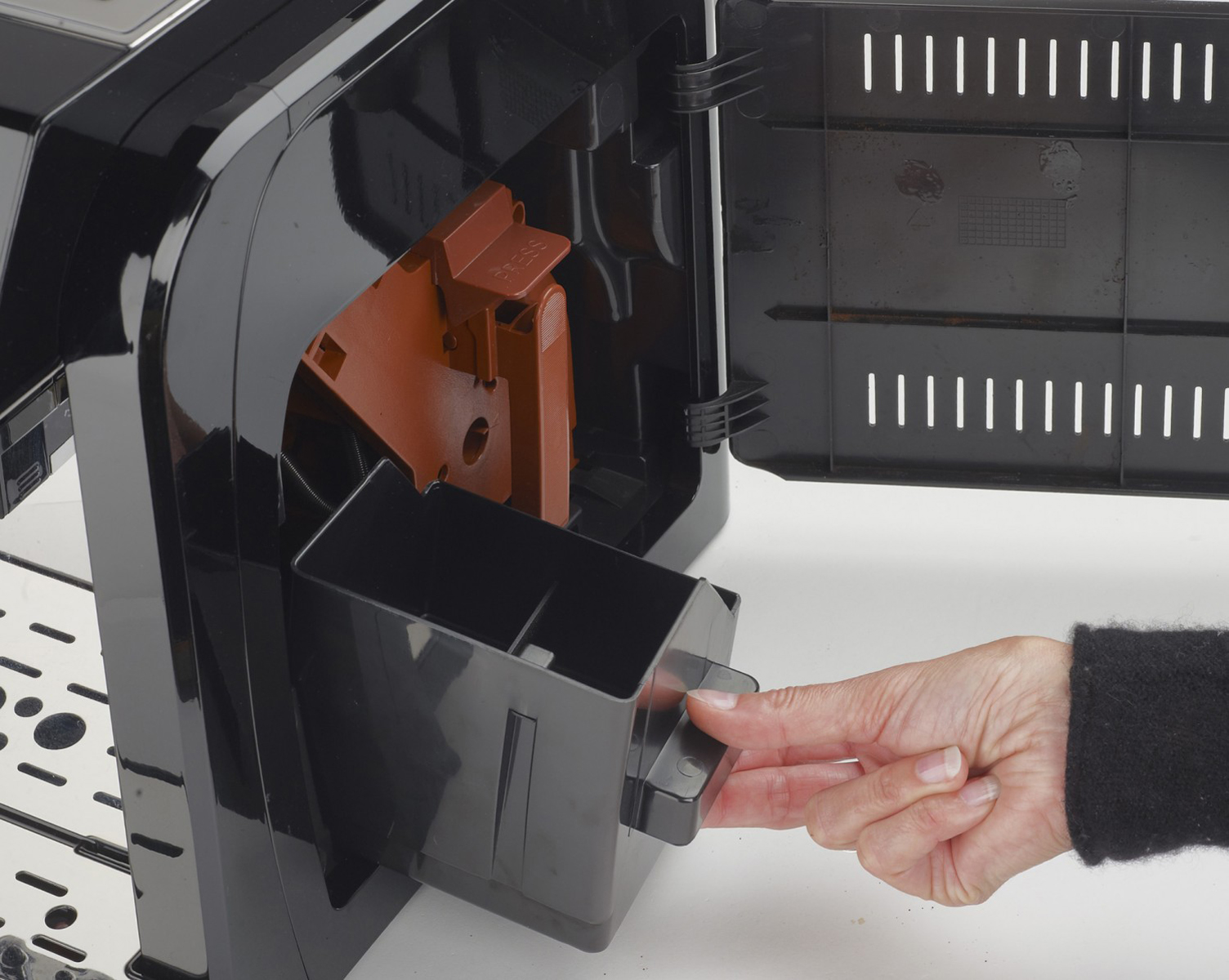 Ręka wyciąga czarną szufladę ekspresu do kawy. Maszyna jest głównie czarna z brązową wkładką.