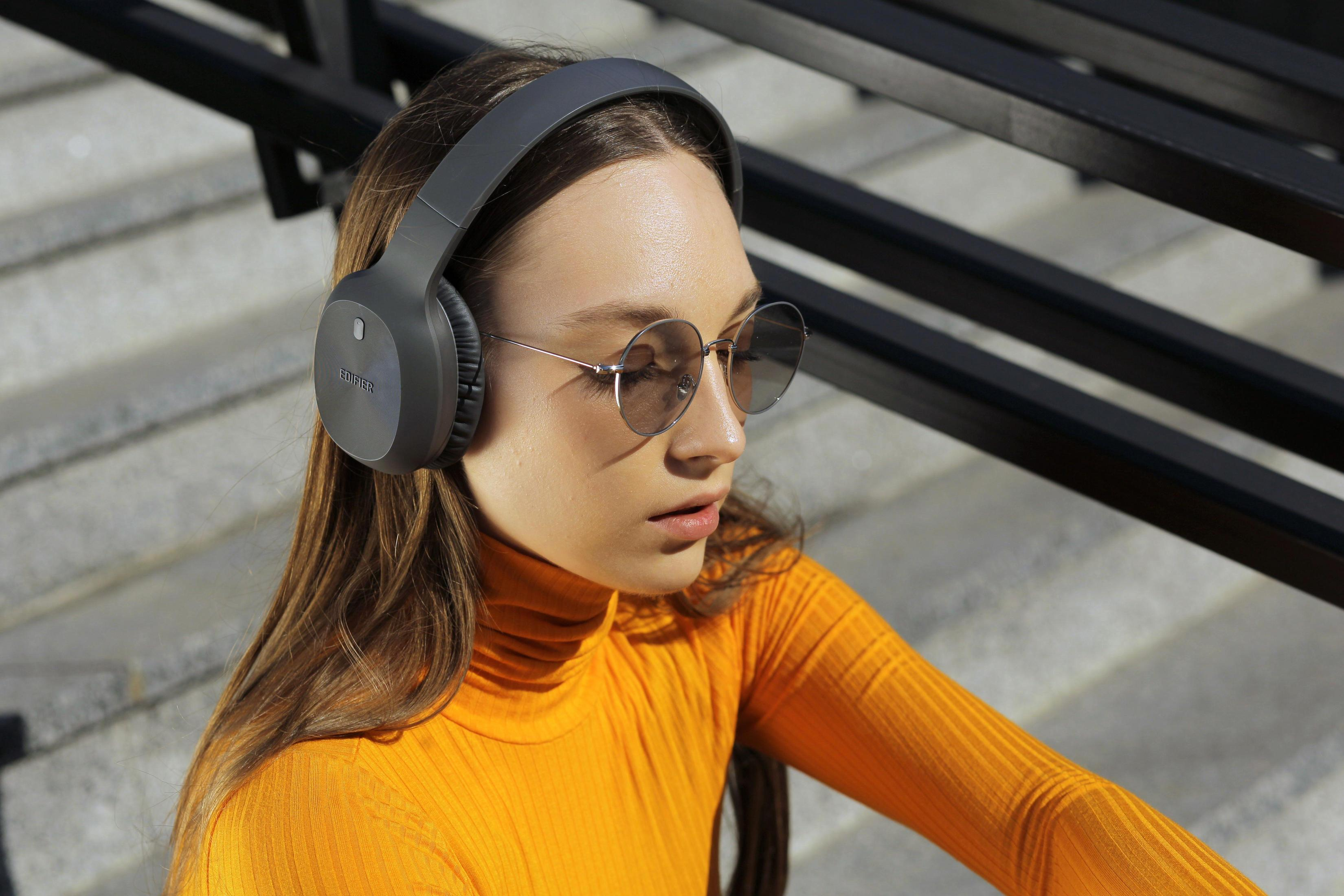 Kobieta w szarych słuchawkach i okularach przeciwsłonecznych. Ma pomarańczową koszulę. Siedzi na zewnątrz.