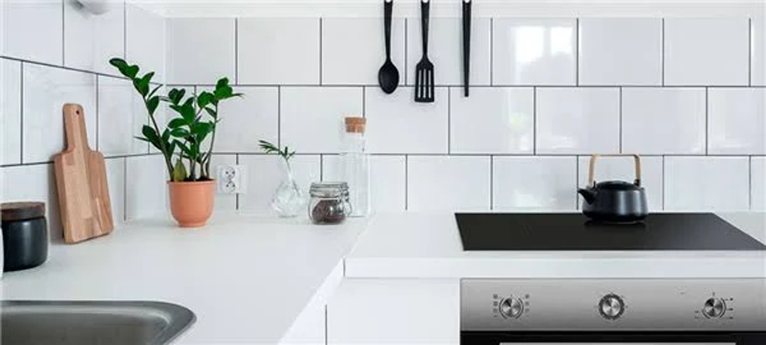 Una cocina blanca con baldosas blancas, una tabla de cortar de madera y una tetera negra.