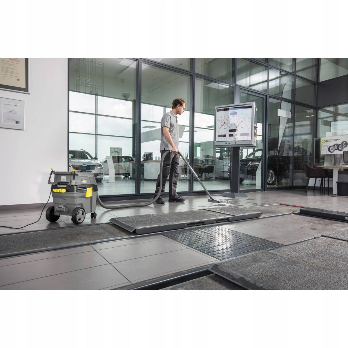 Mężczyzna odkurza podłogę warsztatu samochodowego. Widoczna maszyna czyszcząca.