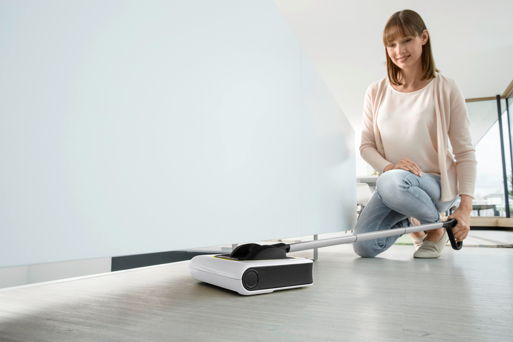Frau benutzt einen Reinigungsroboter auf einem Holzboden. Weiße Wand im Hintergrund.
