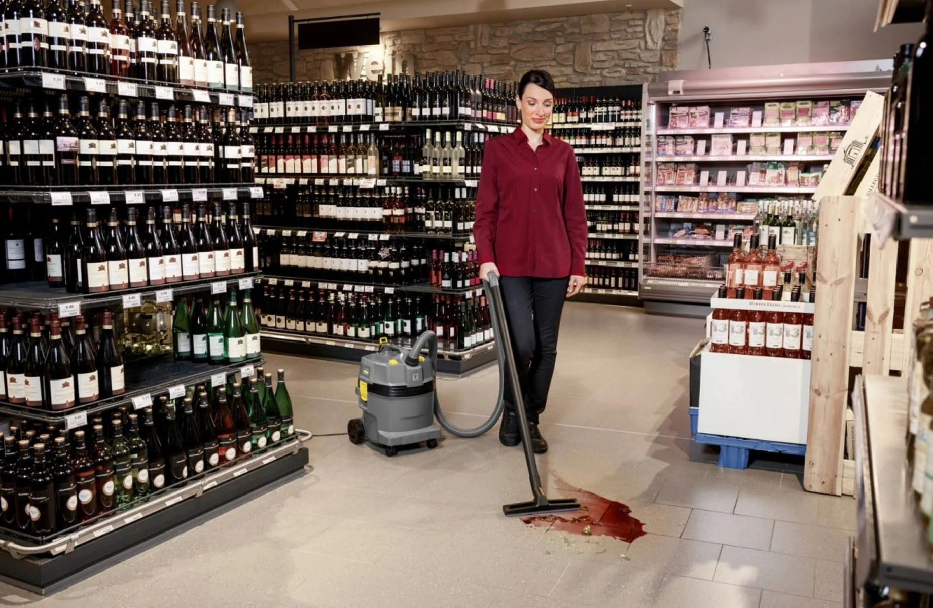 Frau, die eine verschüttete rote Flüssigkeit auf dem Boden in einem Geschäft mit Weinflaschen absaugt.
