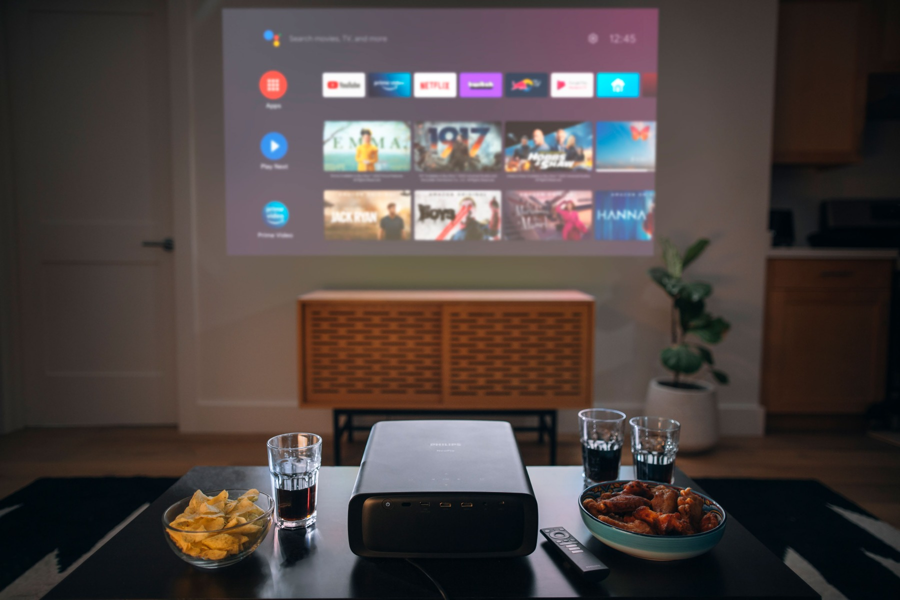 Ein Projektor, der einen Bildschirm mit Streaming-Apps und Filmen projiziert. Snacks und Getränke stehen auf dem Tisch.