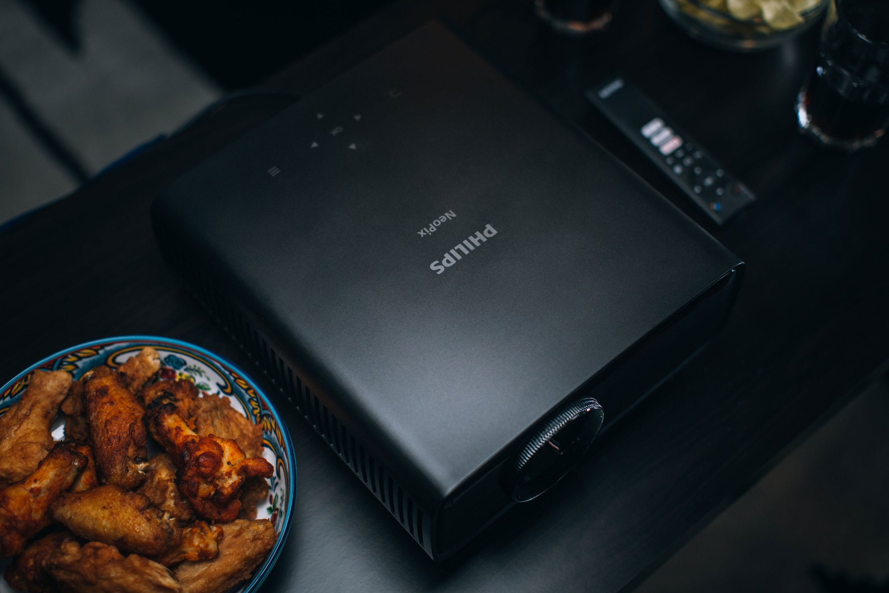 Ein schwarzer Philips NeoPix Projektor auf einem Tisch mit Essen und Fernbedienung. Der Hintergrund ist dunkel.