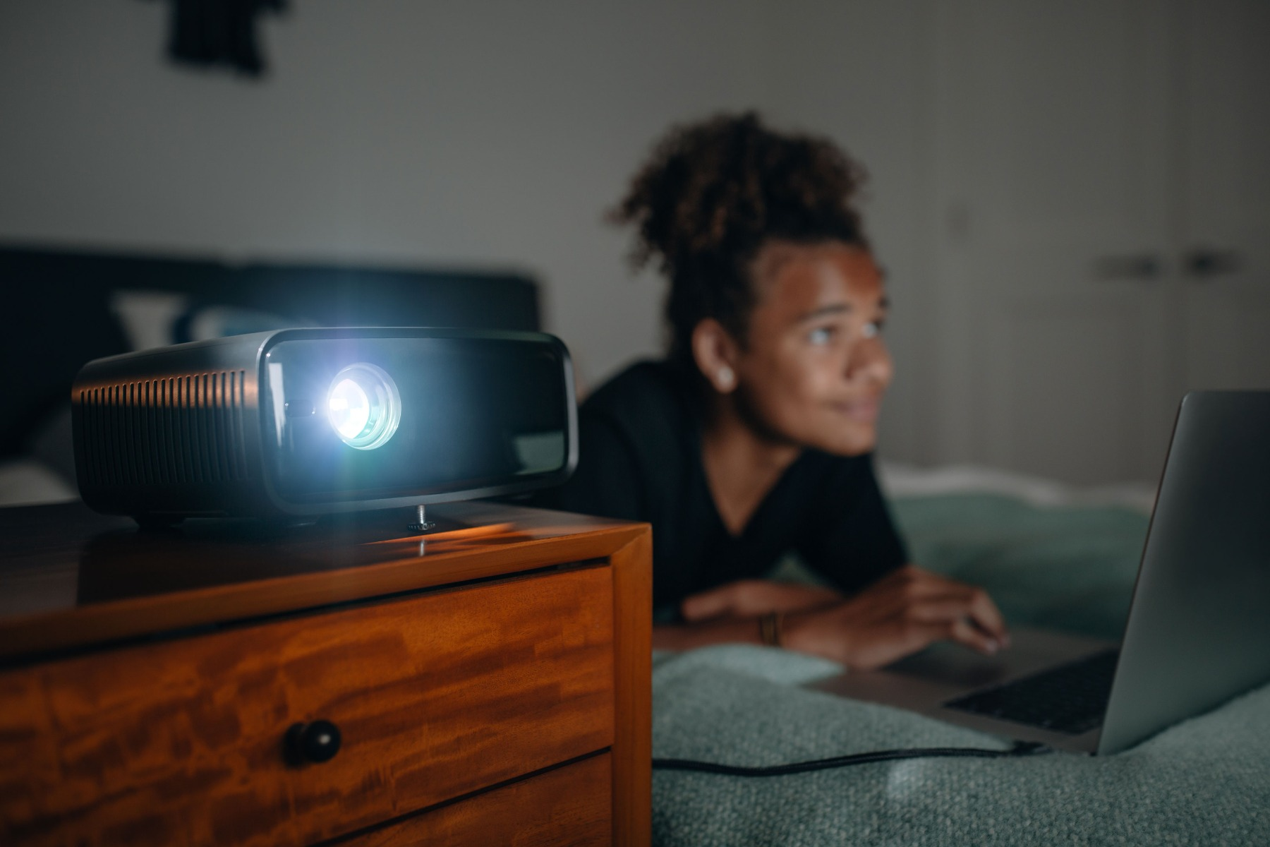 Projektor na szafce nocnej i osoba korzystająca z laptopa.
