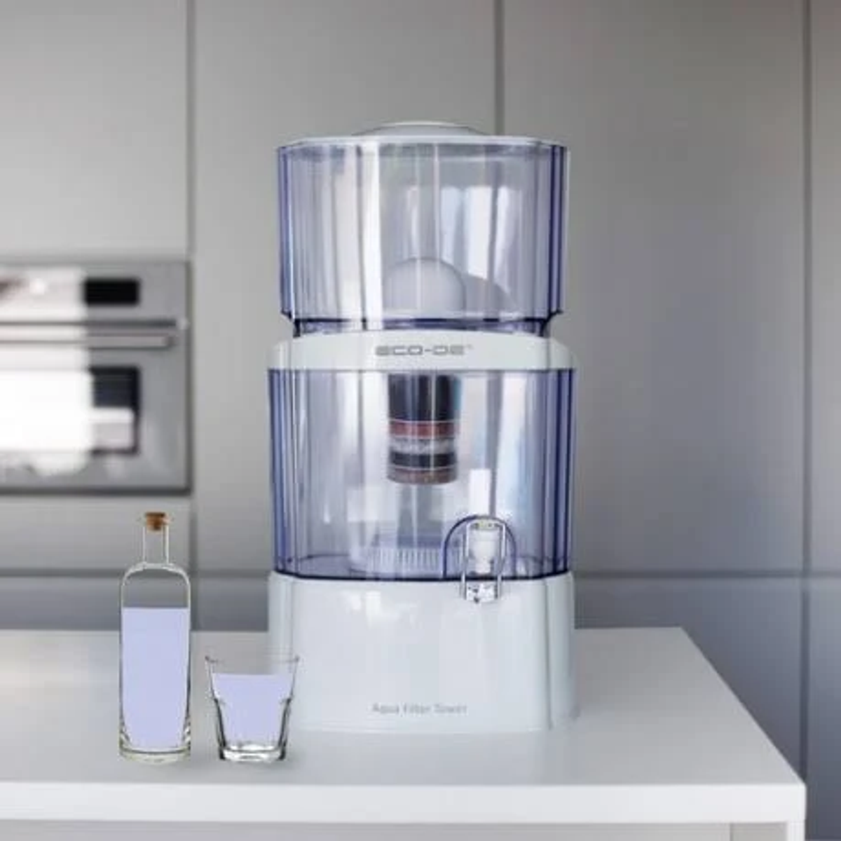 Filtro de agua con una botella y un vaso de agua sobre una encimera blanca en una cocina.