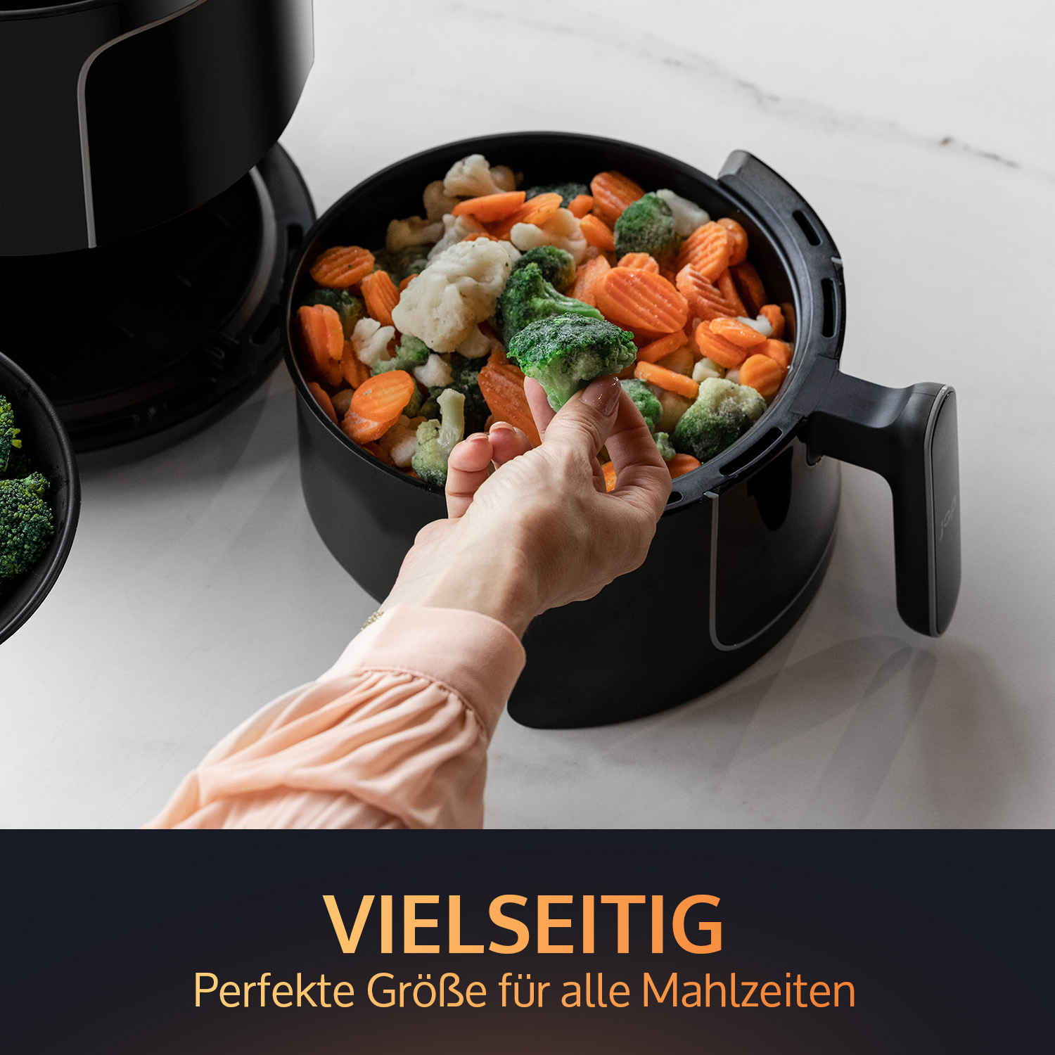 Black air fryer with vegetables. A hand is holding broccoli. The text says "Versatile".