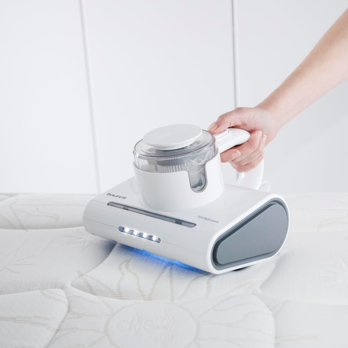 Biały odkurzacz trzymany jest przez dłoń na białym materacu.