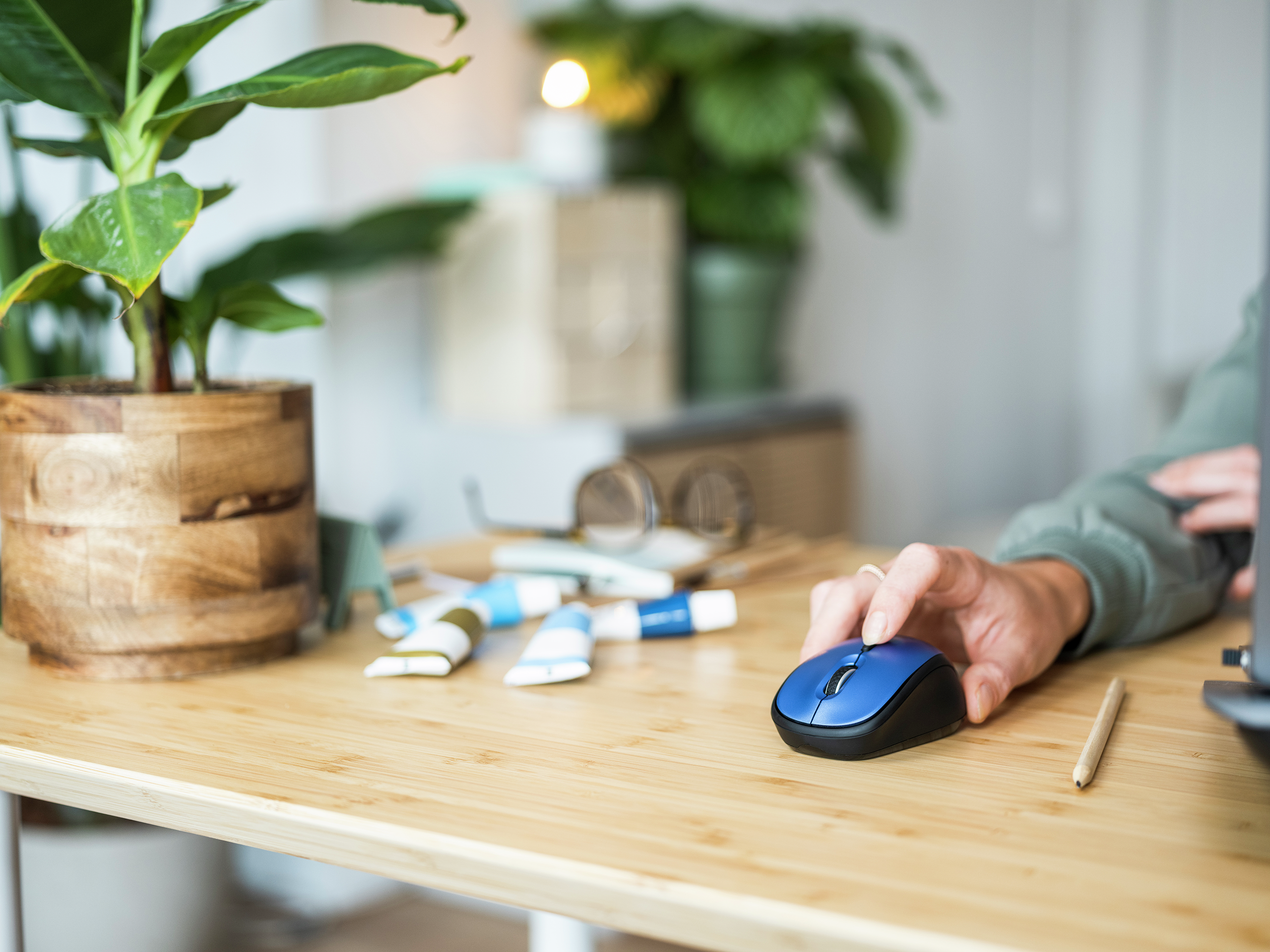 Osoba używająca niebieskiej myszy na drewnianym biurku z przyborami artystycznymi i rośliną.