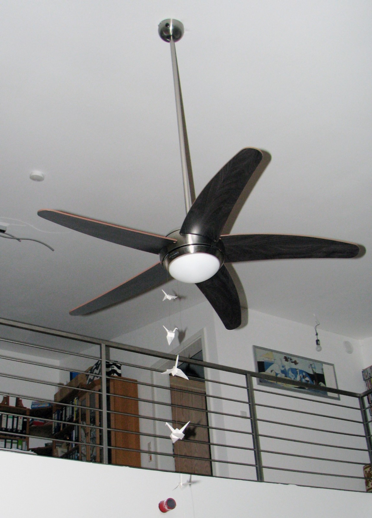 Un ventilatore da soffitto con cinque pale scure e una luce pende da un soffitto bianco. Gru di origami sono sospese sotto.