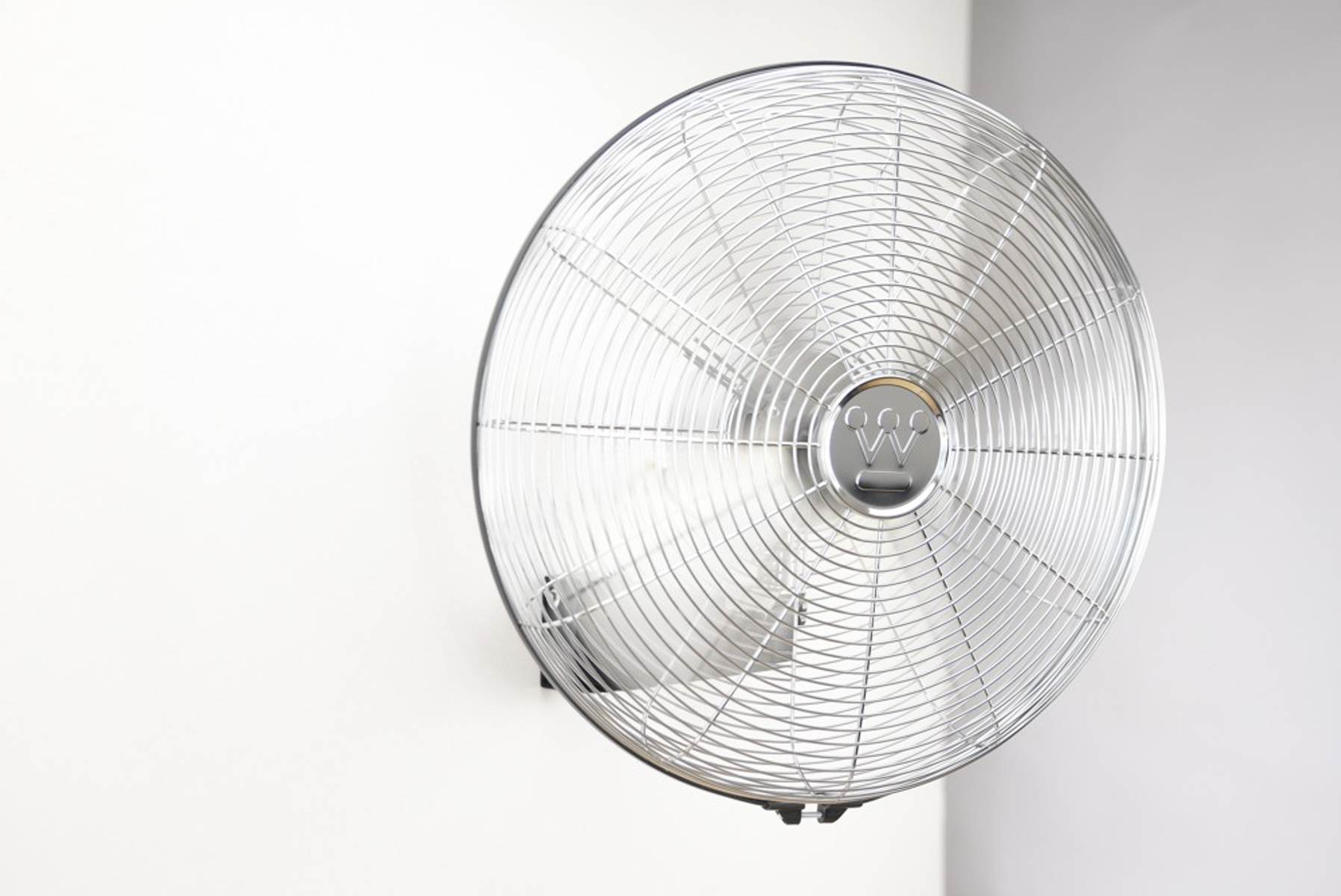 Ein silberner Ventilator an einer weißen Wand. Der Ventilator ist in Bewegung, die Flügel sind sichtbar.