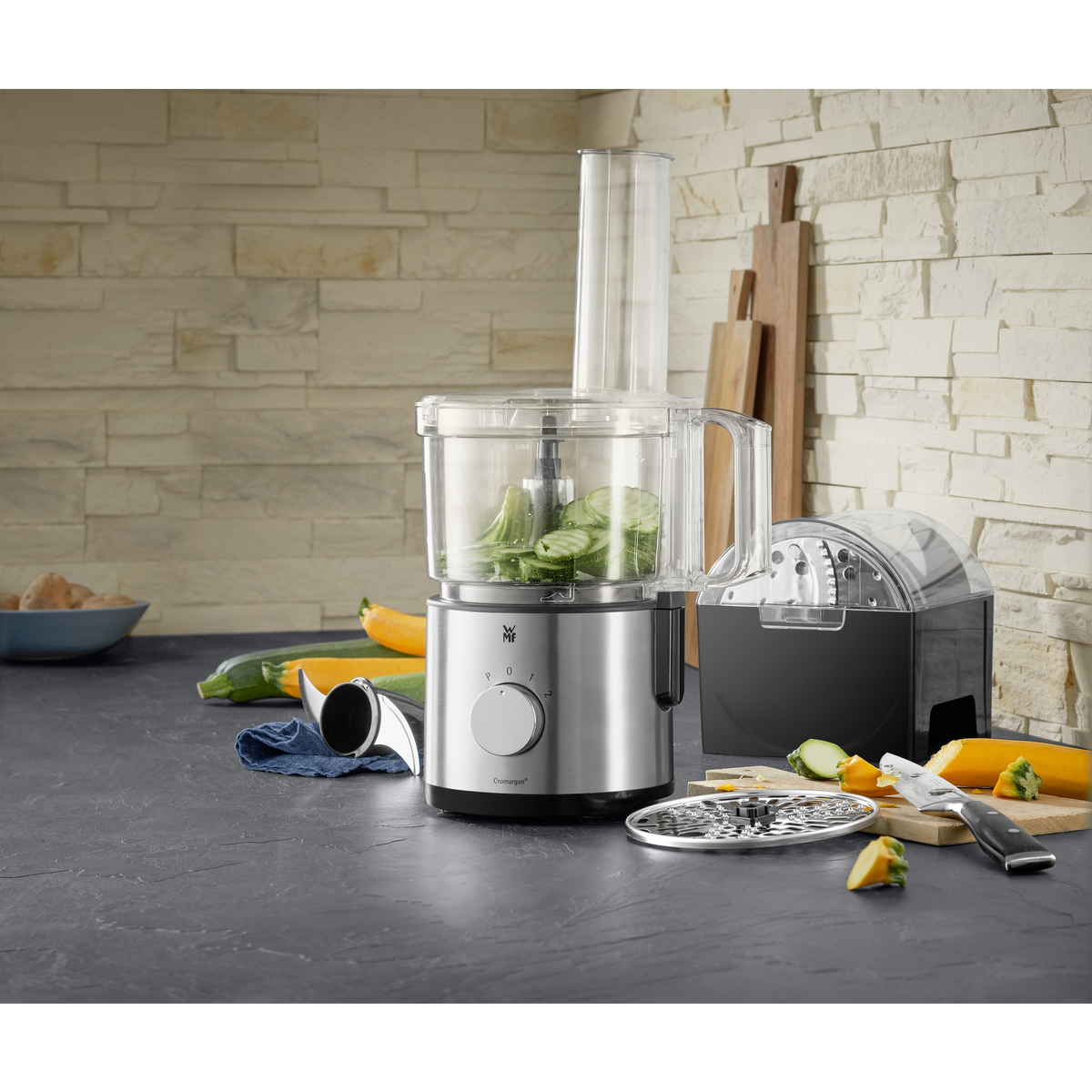 Robot da cucina argentato con verdure, accanto a un'affettatrice nera e taglieri.