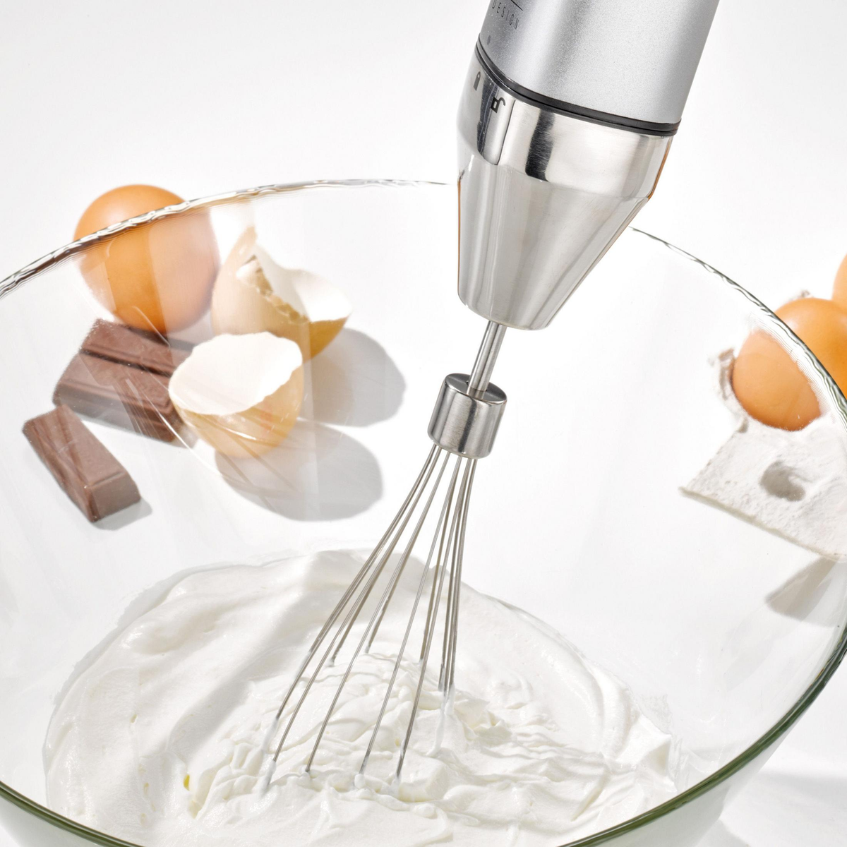 Ein Handmixer, der in einer Schüssel mit weißer Mischung rührt. Eier, zerbrochene Schalen und Schokoladenstücke an der Seite.