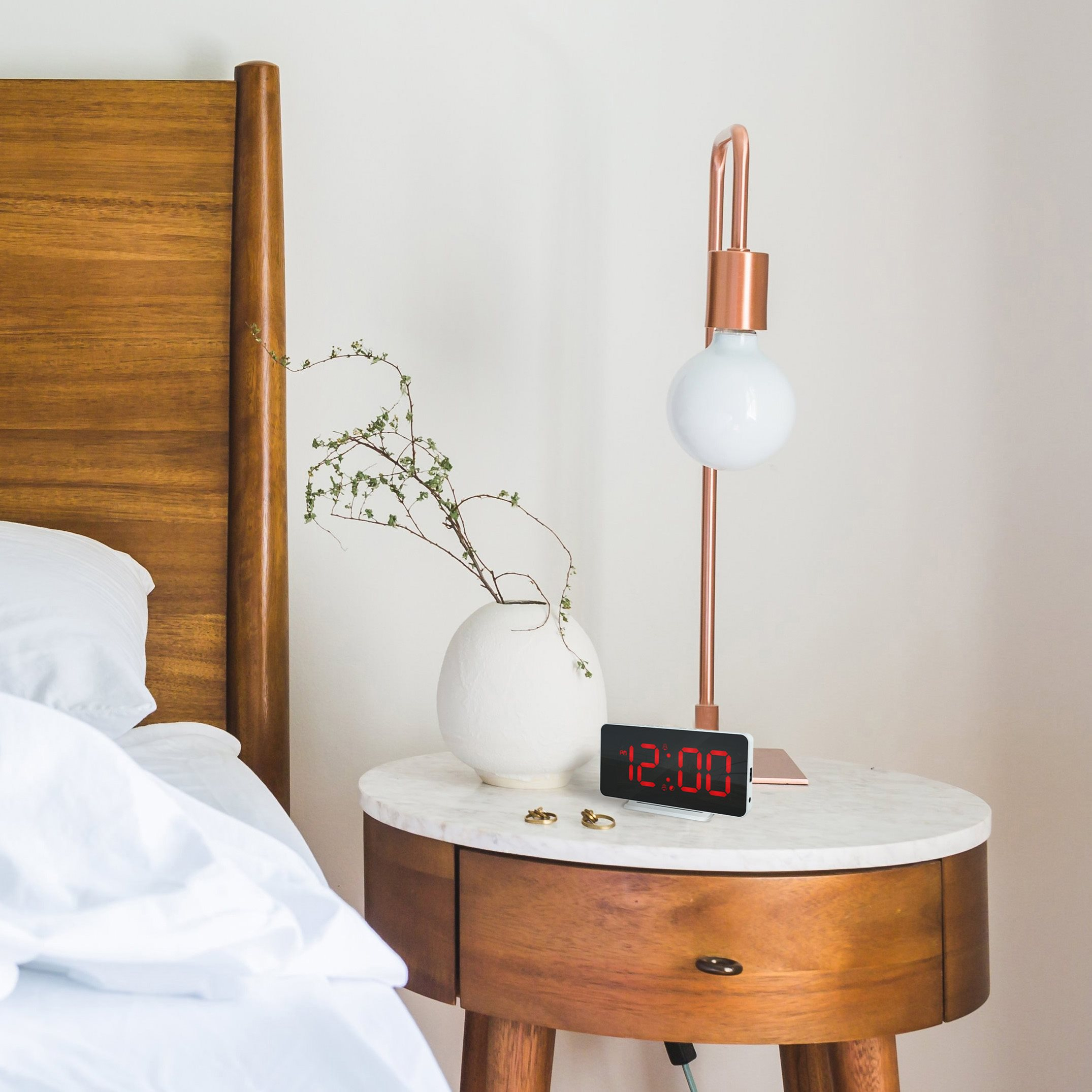 Bedroom scene: bed with white linens, nightstand with lamp, clock, and vase.